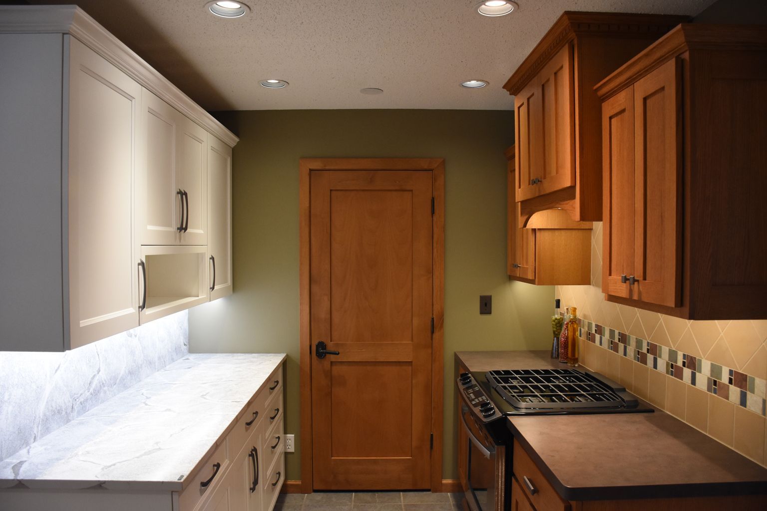 Narrow galley kitchen with white left cabinets, wood right cabinets, marble counters, and a wooden door at the end