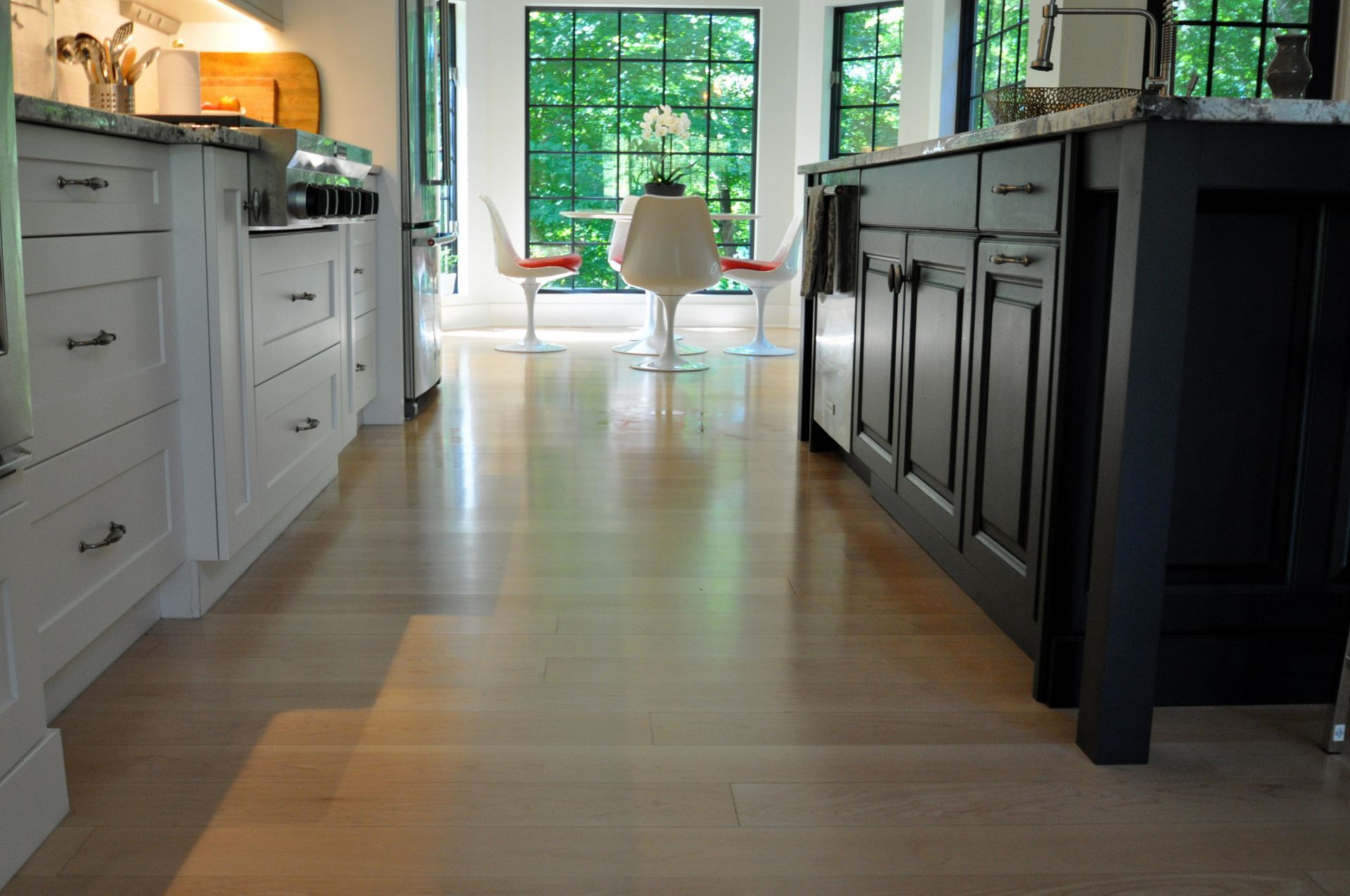 Kitchen hallway with white and dark cabinets, leading to a bright dining area and large window.