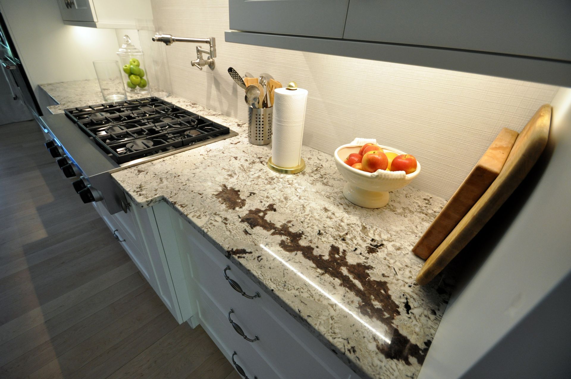 Kitchen countertop with cooktop, speckled granite, fruit bowl, paper towels, and cutting board clutter.