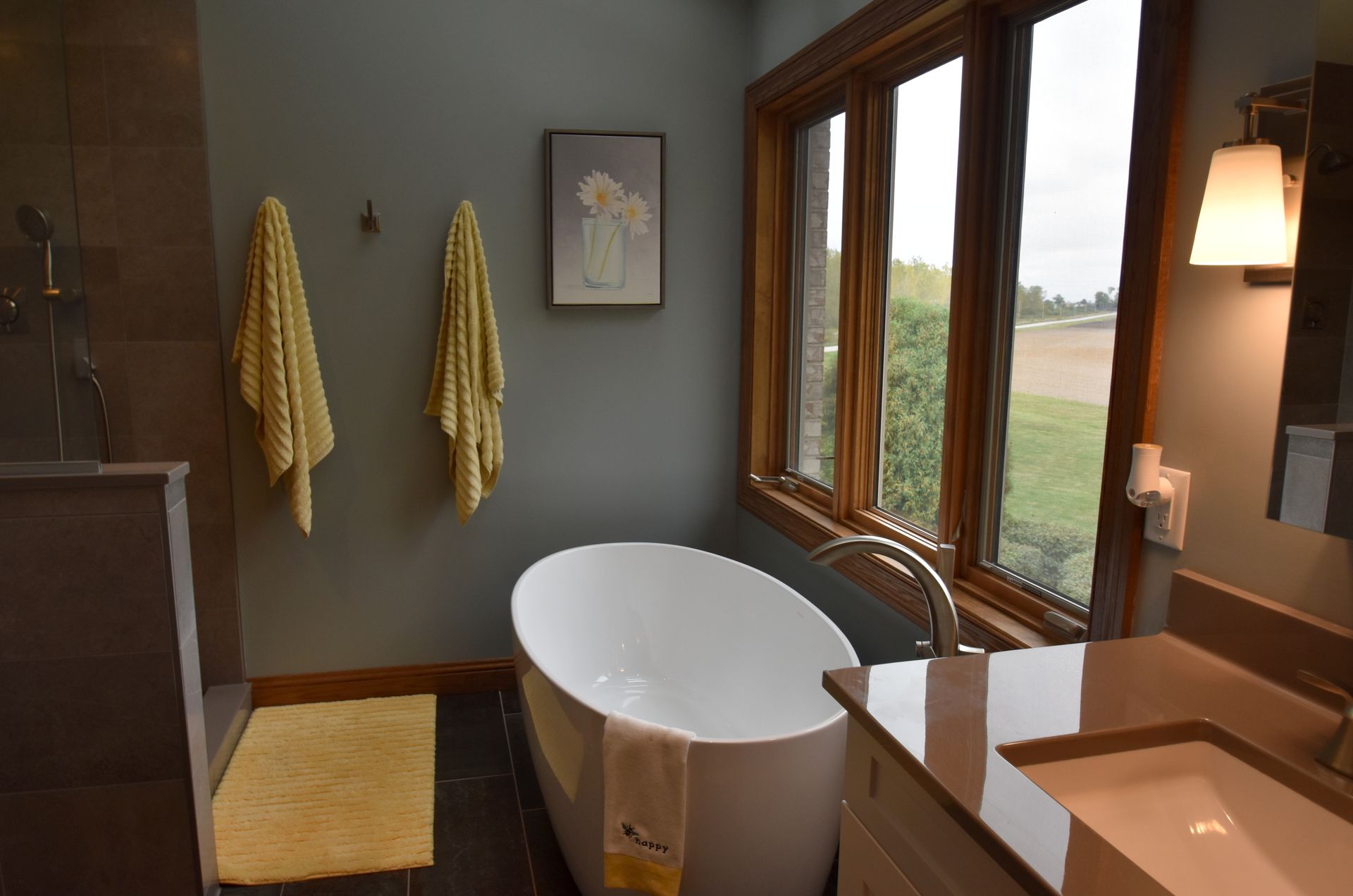 Modern bathroom with freestanding tub, double sinks, large windows, and yellow towels on gray walls