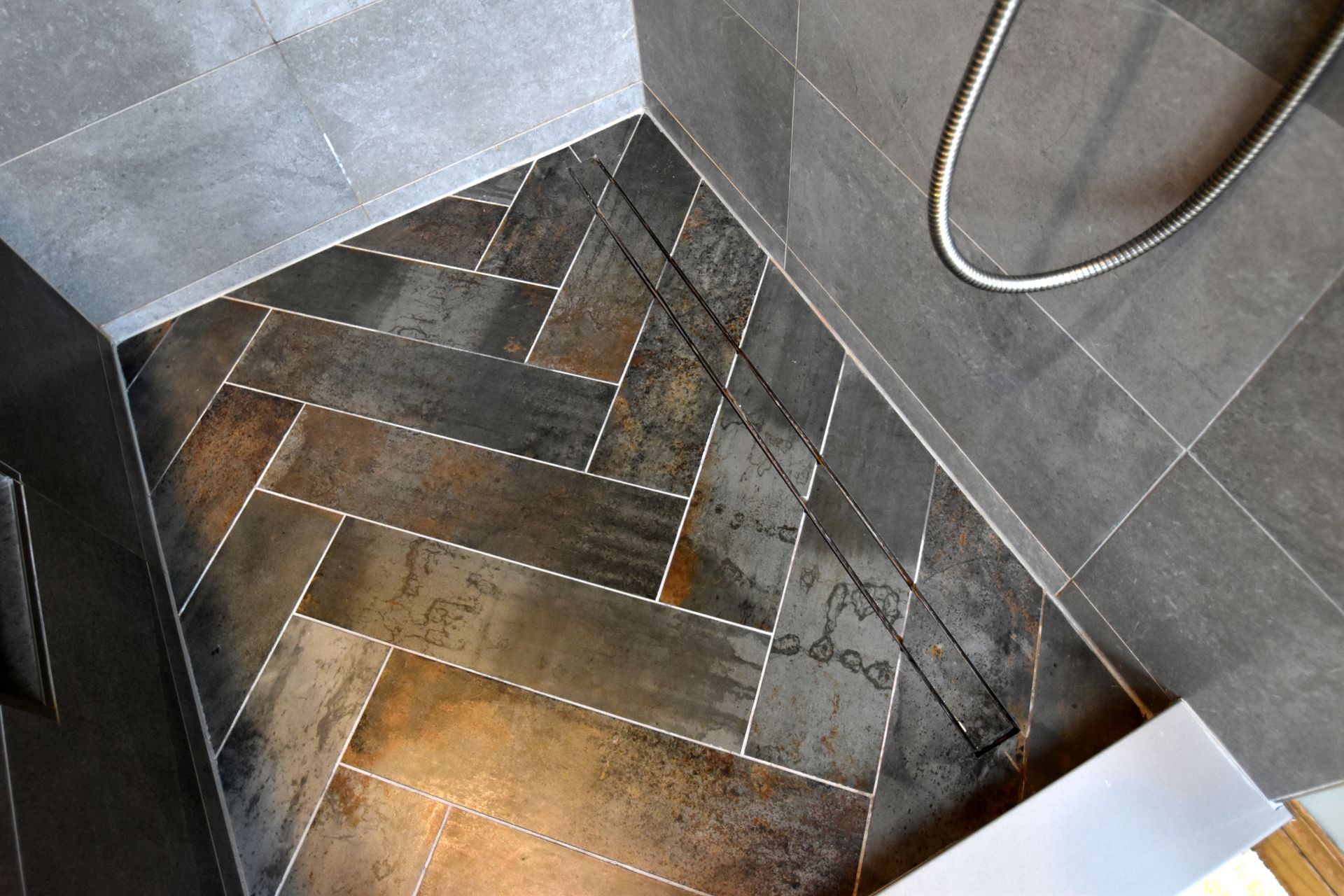 Corner of a tiled shower floor with a drain and a hose against gray walls.