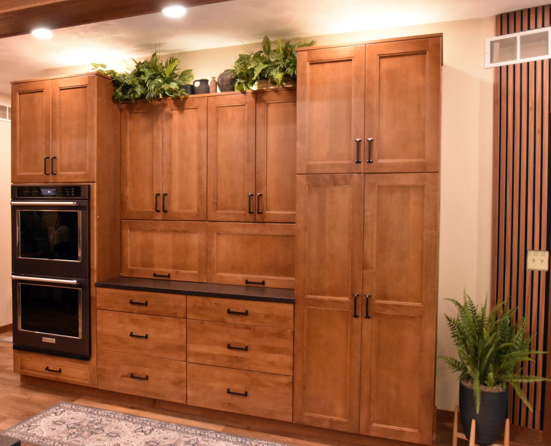 Modern wooden kitchen cabinetry wall with built-in ovens, drawers, and potted plants on top and beside it