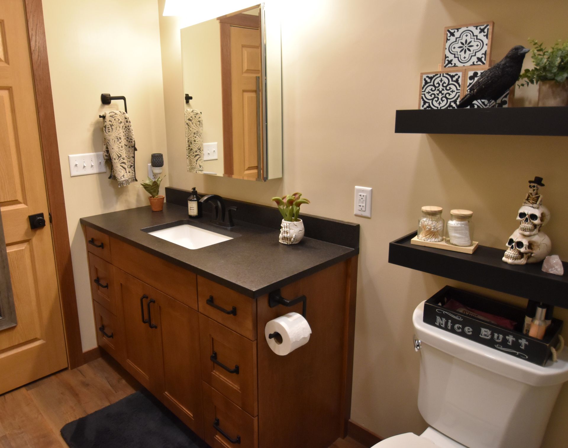 Bathroom vanity with black countertop, mirror, towel ring, and toilet paper holder beside a toilet and wall shelves