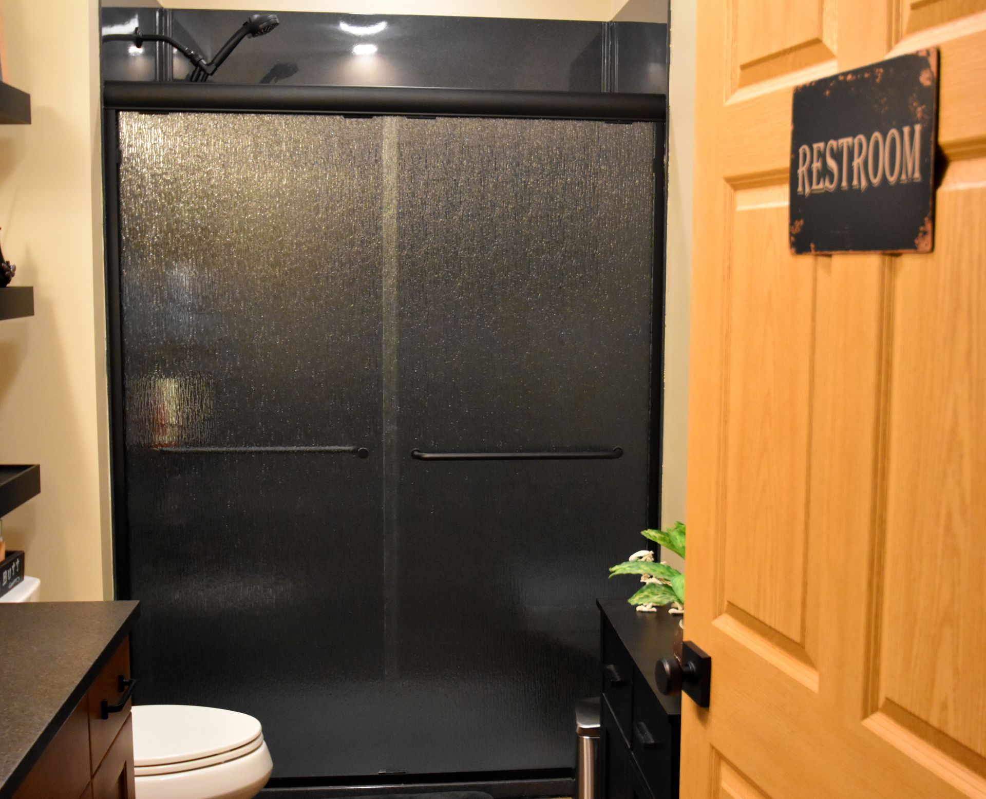 Bathroom shower with black sliding doors and a wooden door labeled “RESTROOM” nearby.