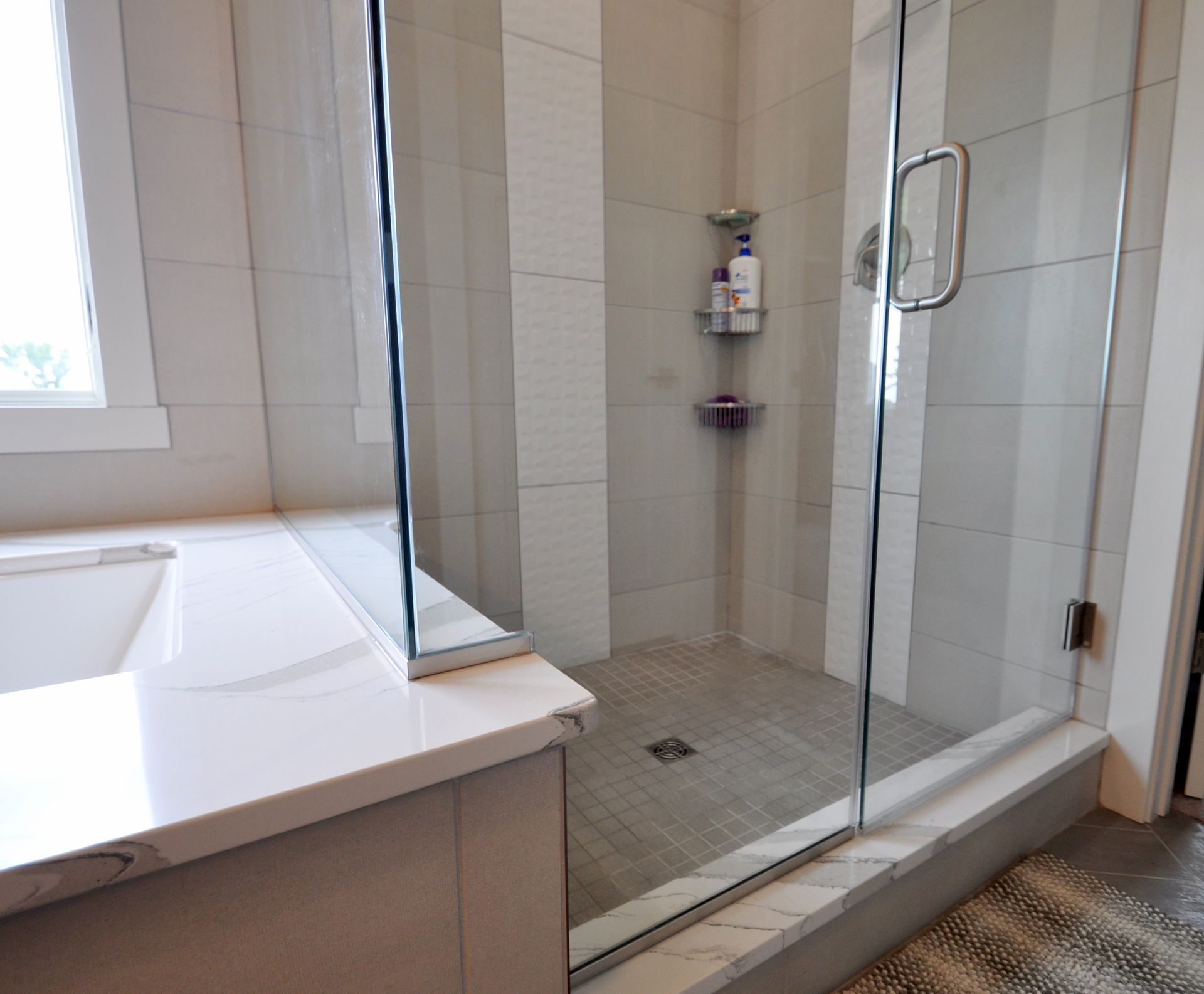 Modern bathroom with glass-enclosed shower, white tile, and bathtub beside a window