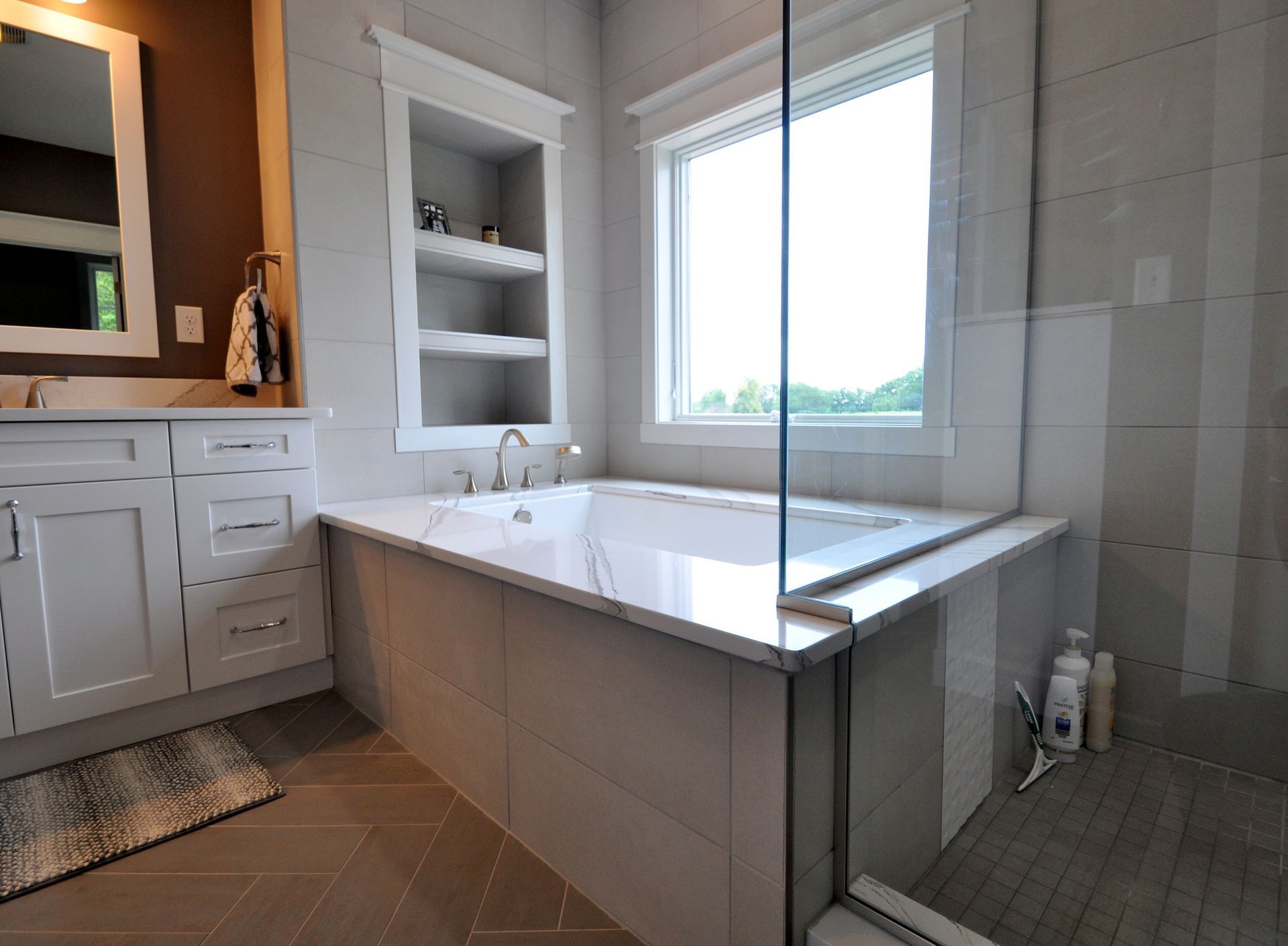 Modern bathroom with a freestanding tub, white vanity, mirror, and tiled floor, viewed through a glass shower panel