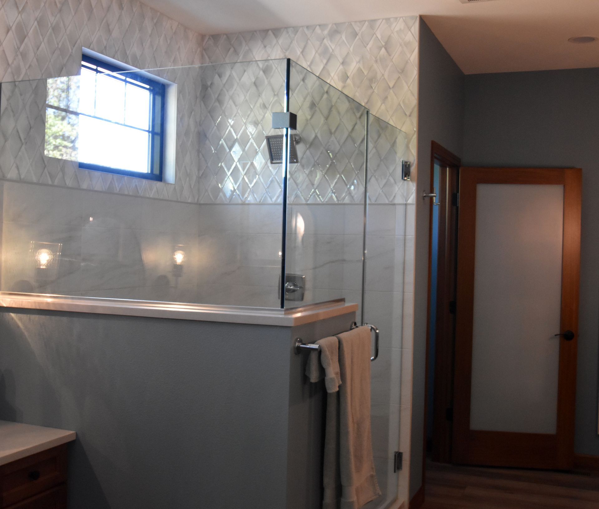 Modern bathroom with glass shower, gray tile, frosted window, and towels hanging on the wall.