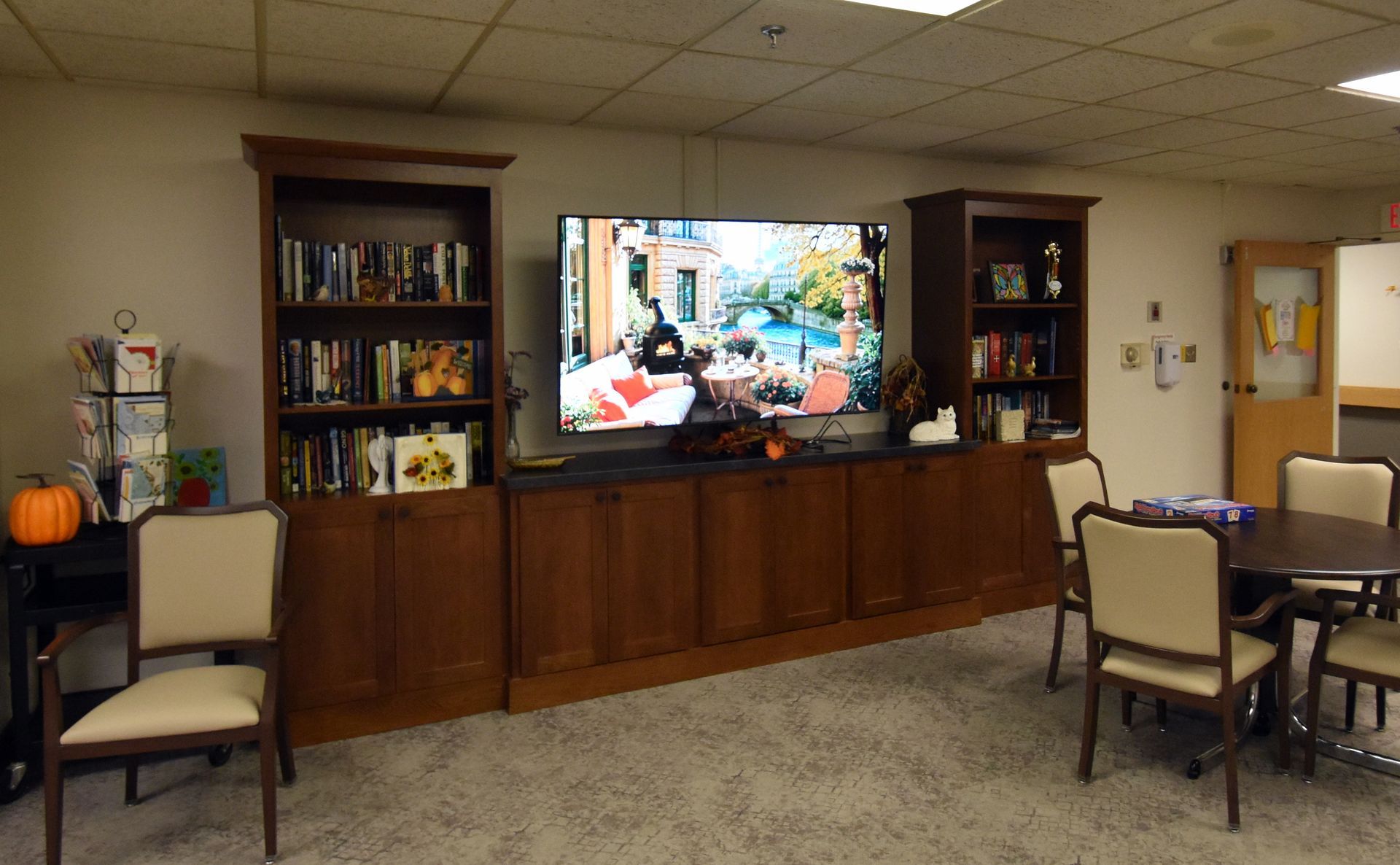 Lounge with a large TV centered in a wooden cabinet, shelves, and small seating area with tables and chairs.