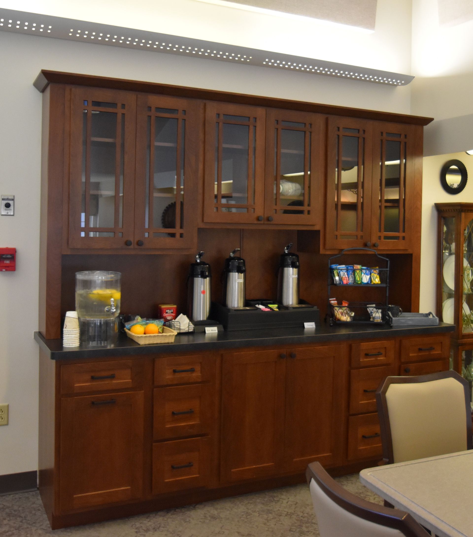 Wooden kitchen cabinet with coffee and drink dispensers on a dark countertop in an office break room.