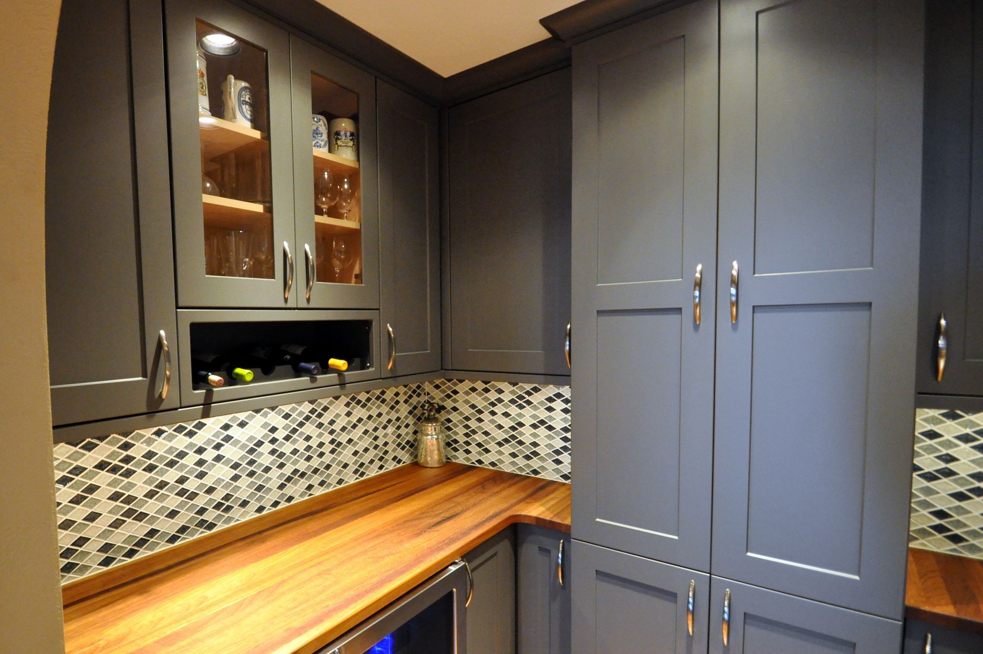 Corner kitchen with dark gray cabinets, wood countertop, mosaic backsplash, and built-in wine cooler.