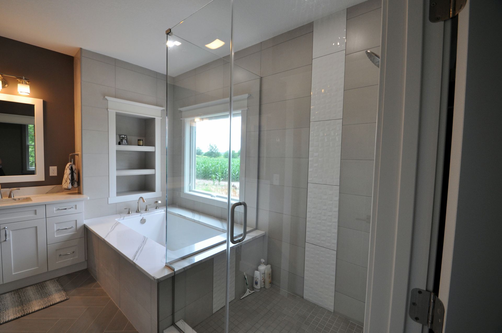 Bright modern bathroom with glass shower, white tub, vanity, and large window overlooking greenery