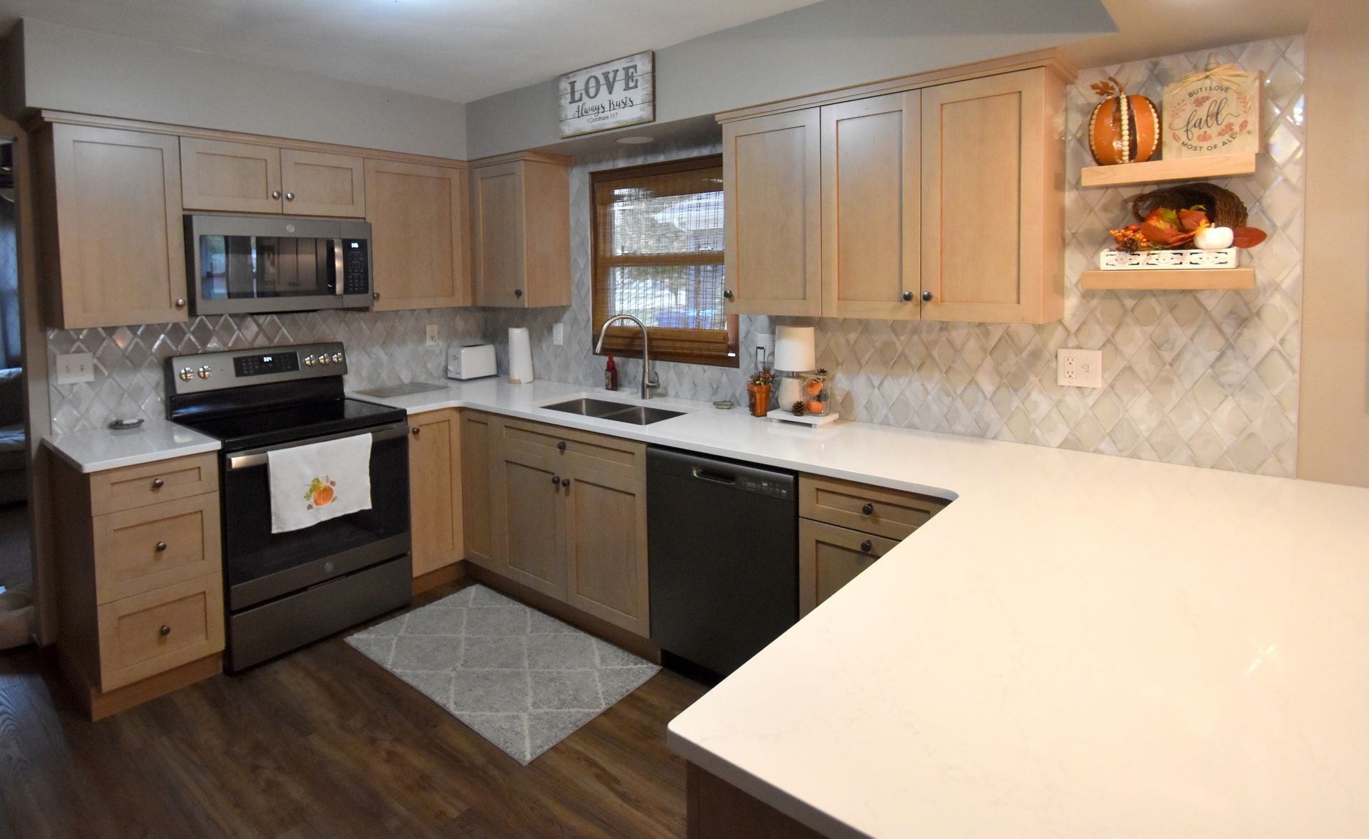 Modern kitchen with light wood cabinets, black appliances, beige counters, and a window over the sink.