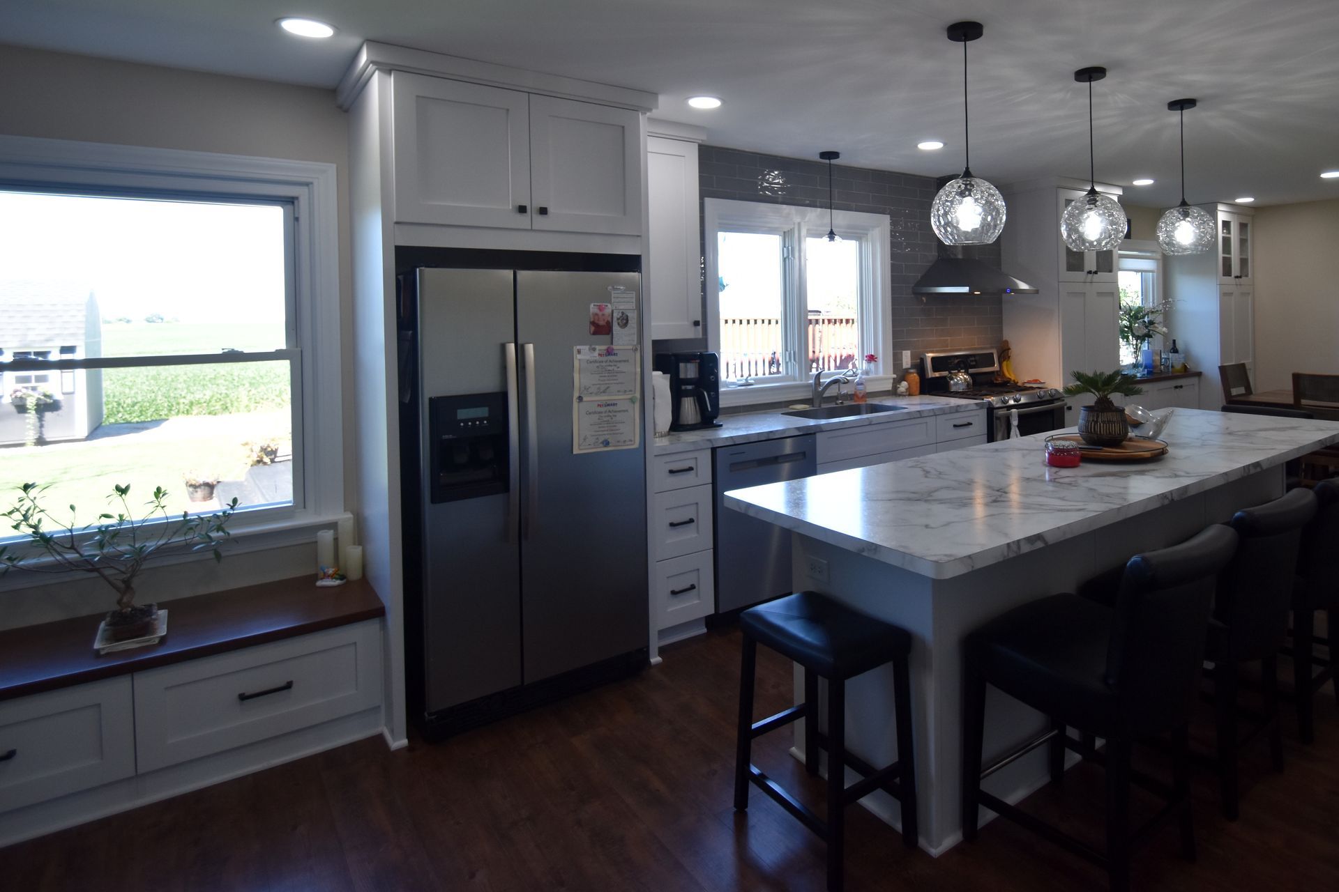 Modern kitchen with large island, stainless steel fridge, pendant lights, and a bright window seat.