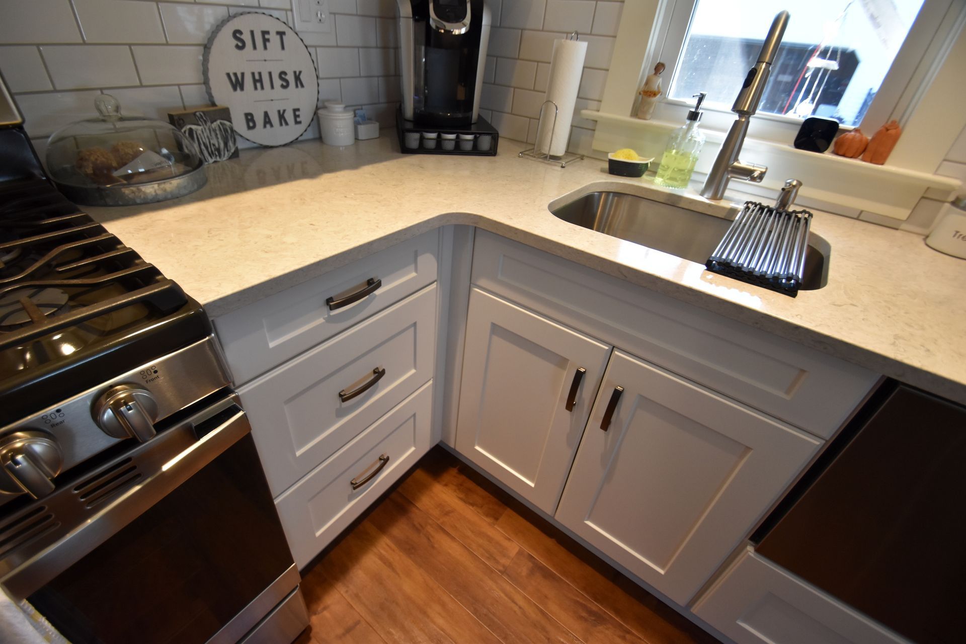 Modern kitchen corner with white cabinets, stainless stove, wood floor, and a window over the sink.