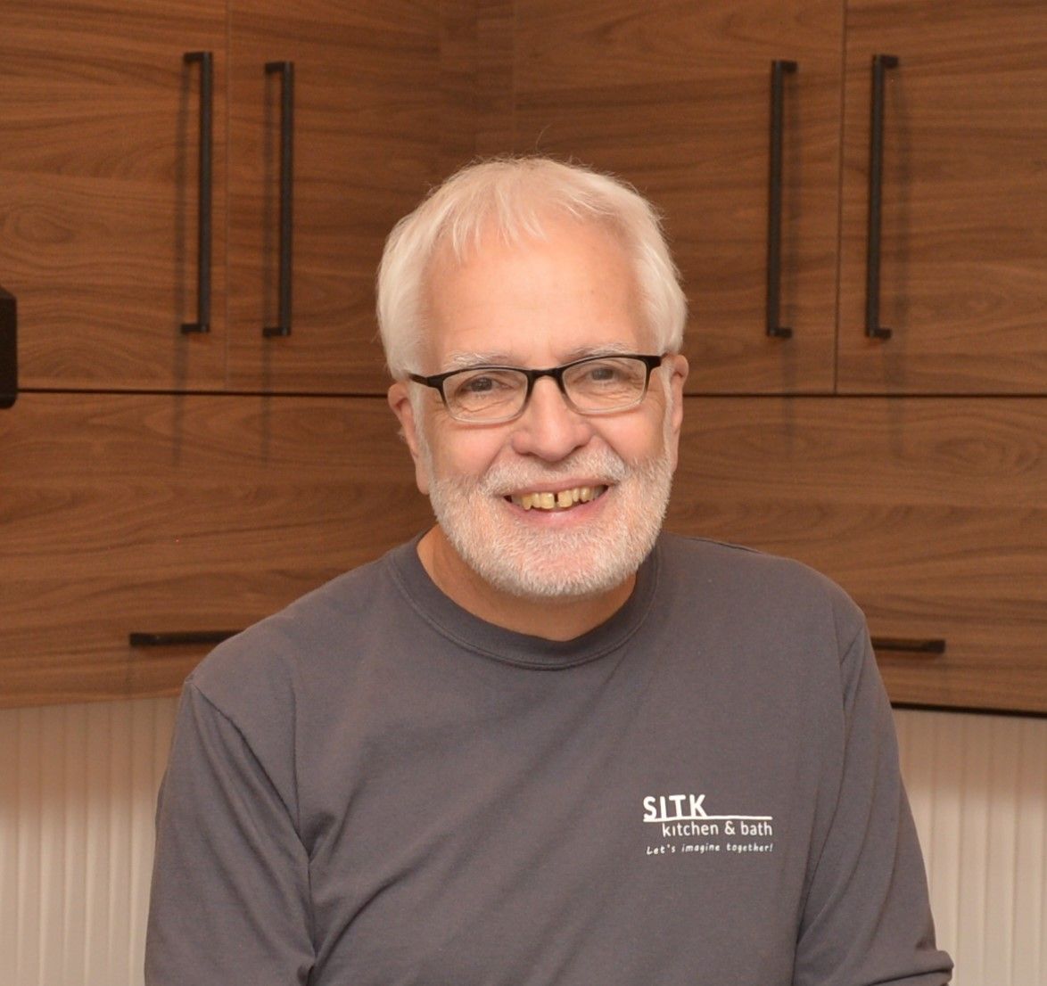 Smiling man with white hair and glasses in a gray shirt standing in a kitchen