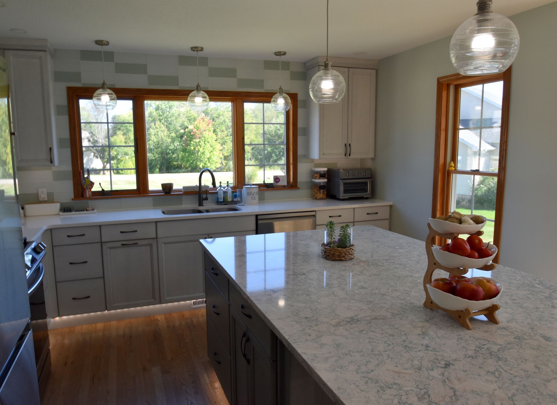 Modern kitchen with gray cabinets, white island, pendant lights, and large window over the sink.