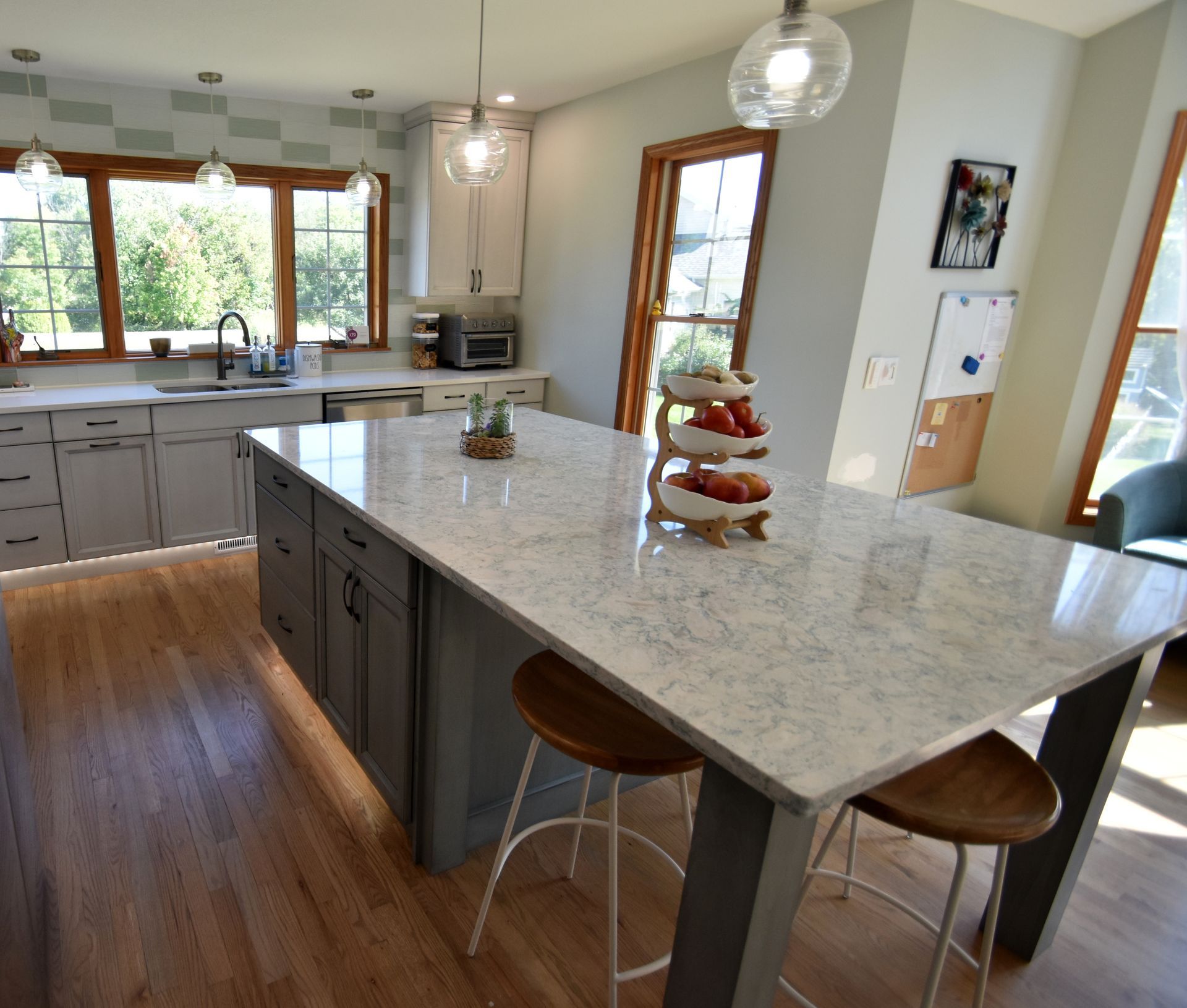 Bright kitchen with a large marble island, wooden floors, and windows overlooking greenery