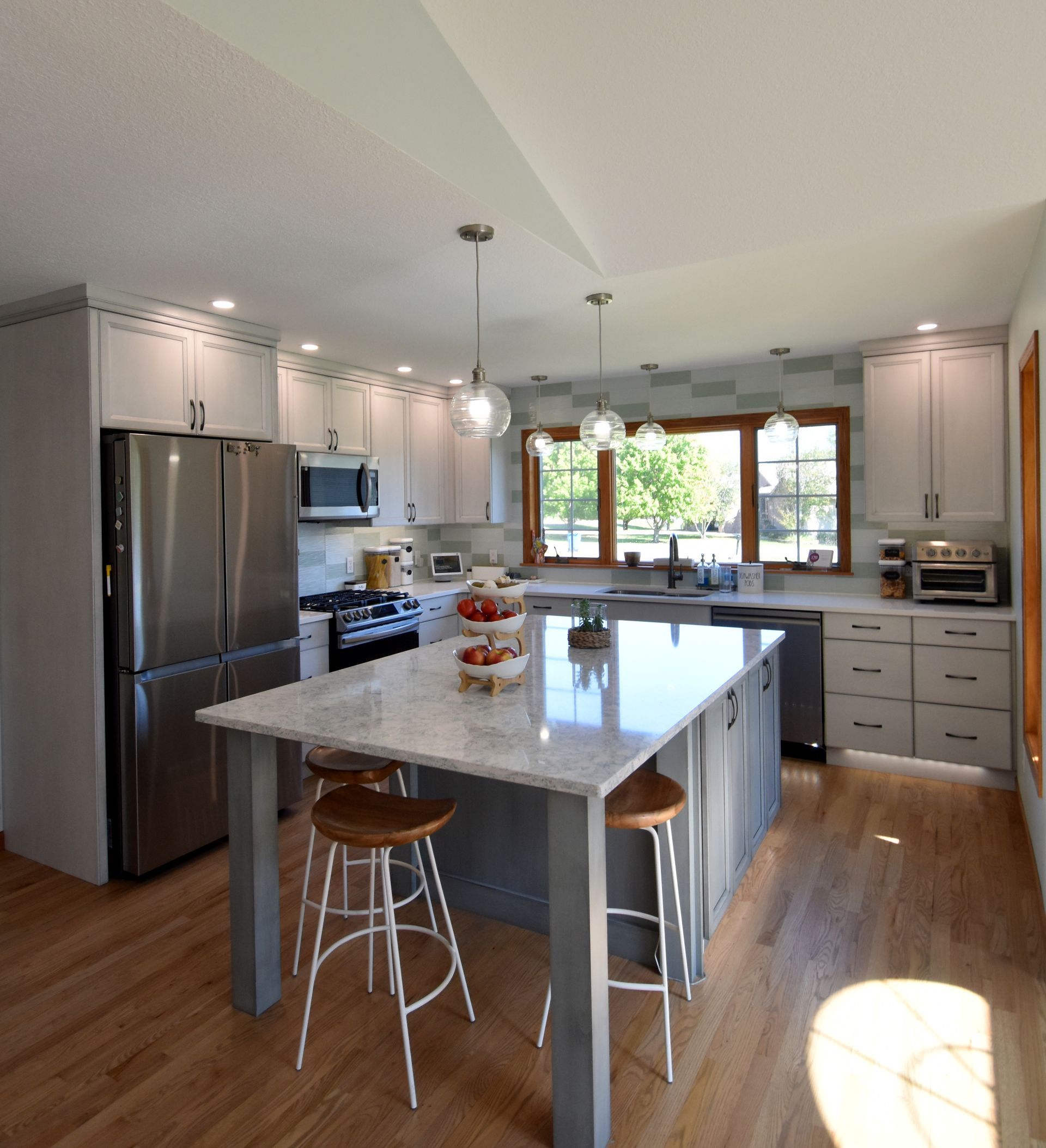 Bright modern kitchen with stainless fridge, island, white cabinets, and large window overlooking greenery