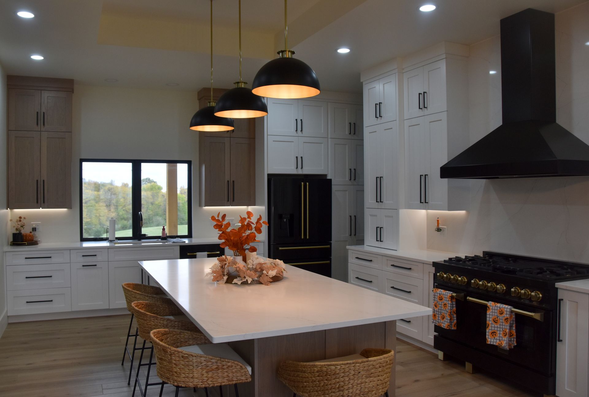 Modern white kitchen with island, black pendant lights, and a large window showing greenery.