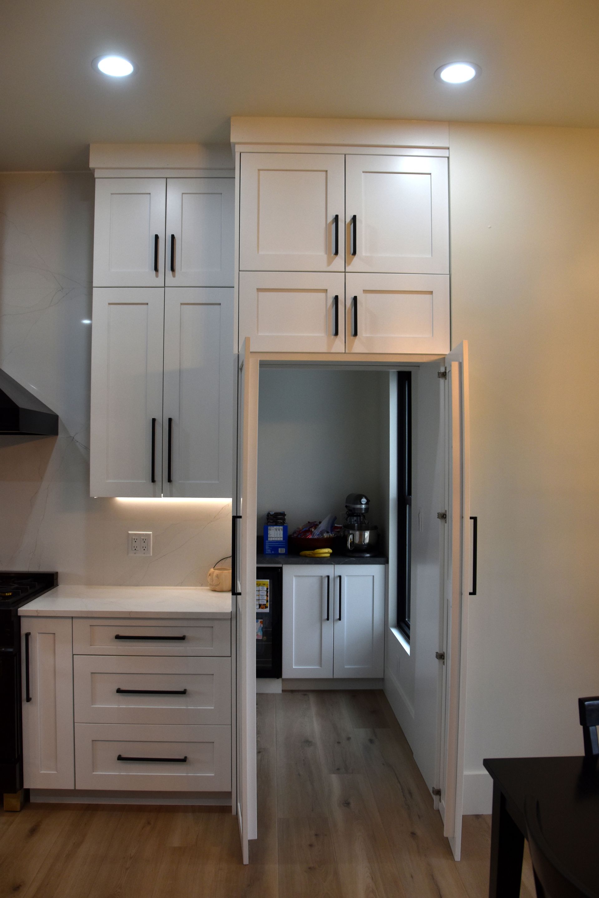Small white cabinets and drawers in a kitchen hallway with recessed ceiling lights and open doorway.