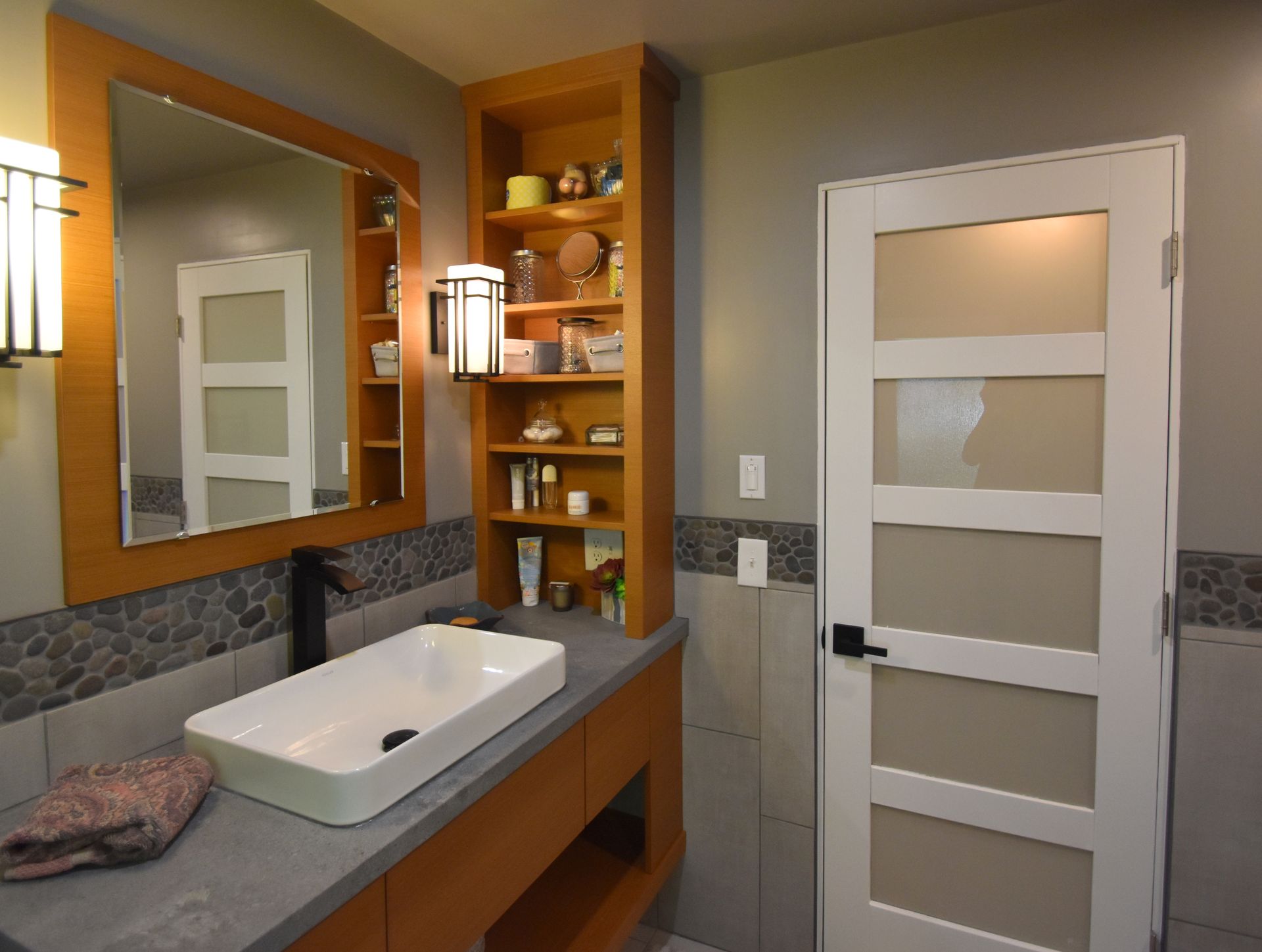 Modern bathroom with white sink, large mirror, wood shelving, and frosted glass door