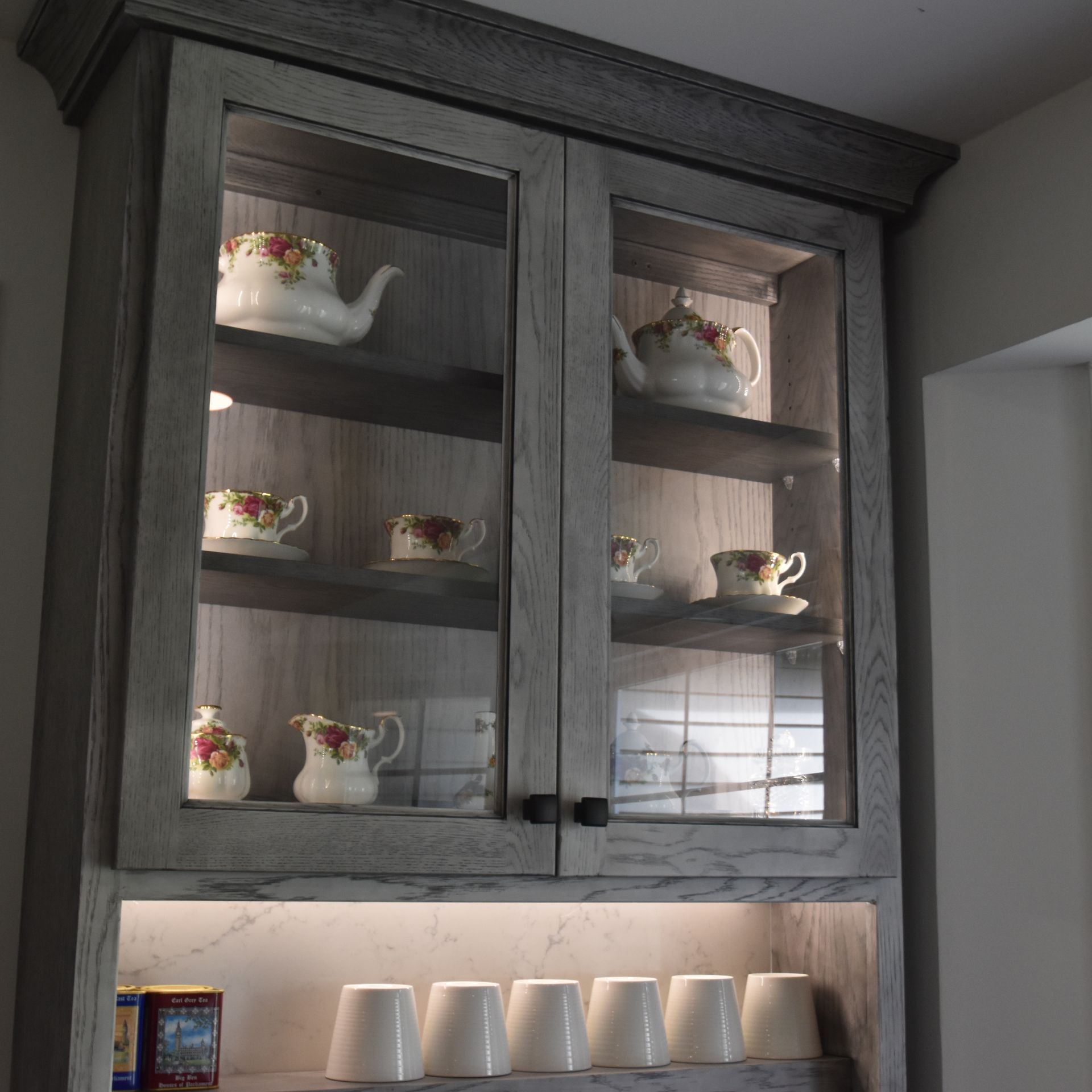 Gray kitchen cabinet with glass doors displaying floral teapots and cups, lit from below.