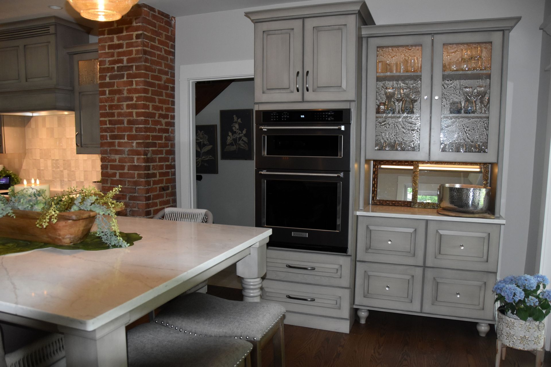 Modern kitchen with gray cabinets, brick accent wall, island, and built-in oven and glass-front hutch