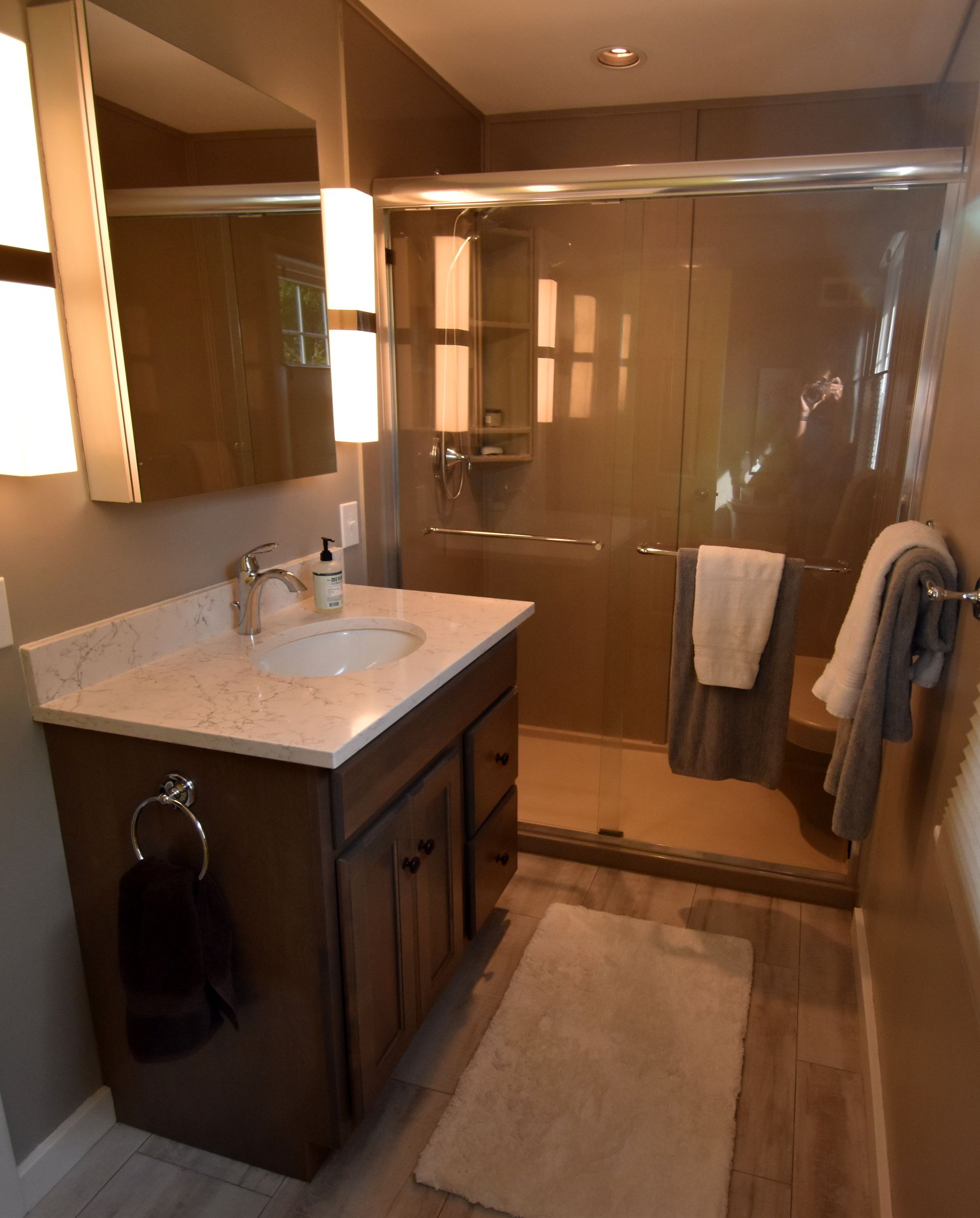 Modern bathroom with wood vanity, large mirror, glass shower, and beige floor mat.