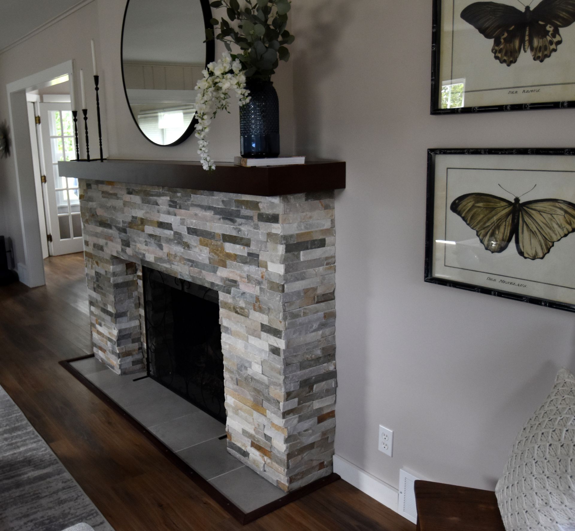 Living room fireplace with gray stone surround, dark wood mantel, and butterfly wall art