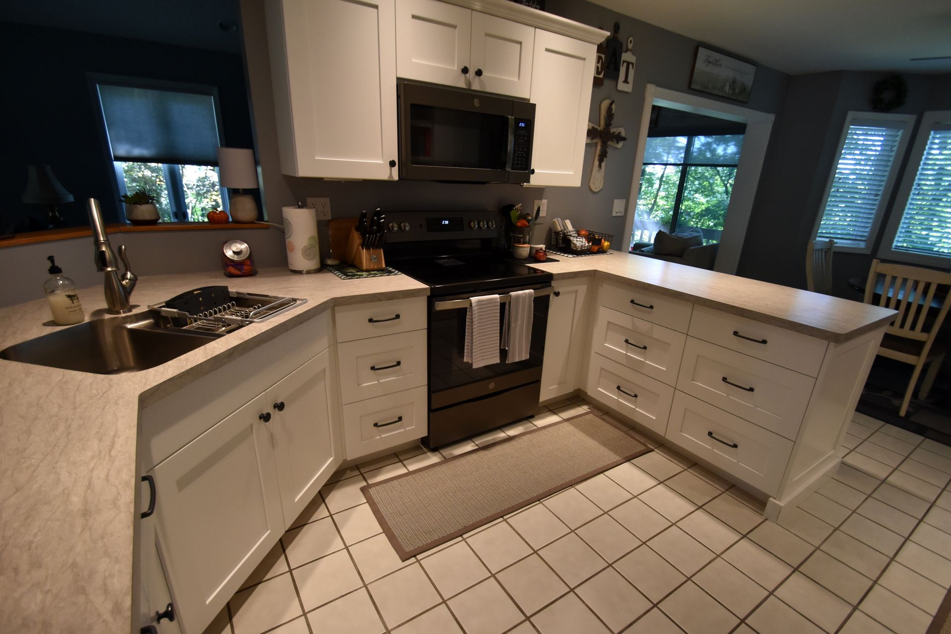Bright kitchen with white cabinets, black stove and sink, tiled floor, and large windows overlooking greenery