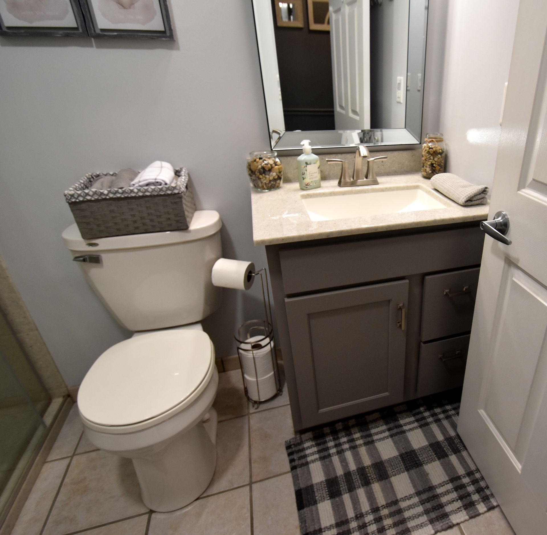 Small bathroom with white toilet, dark vanity, mirror, and plaid floor rug.