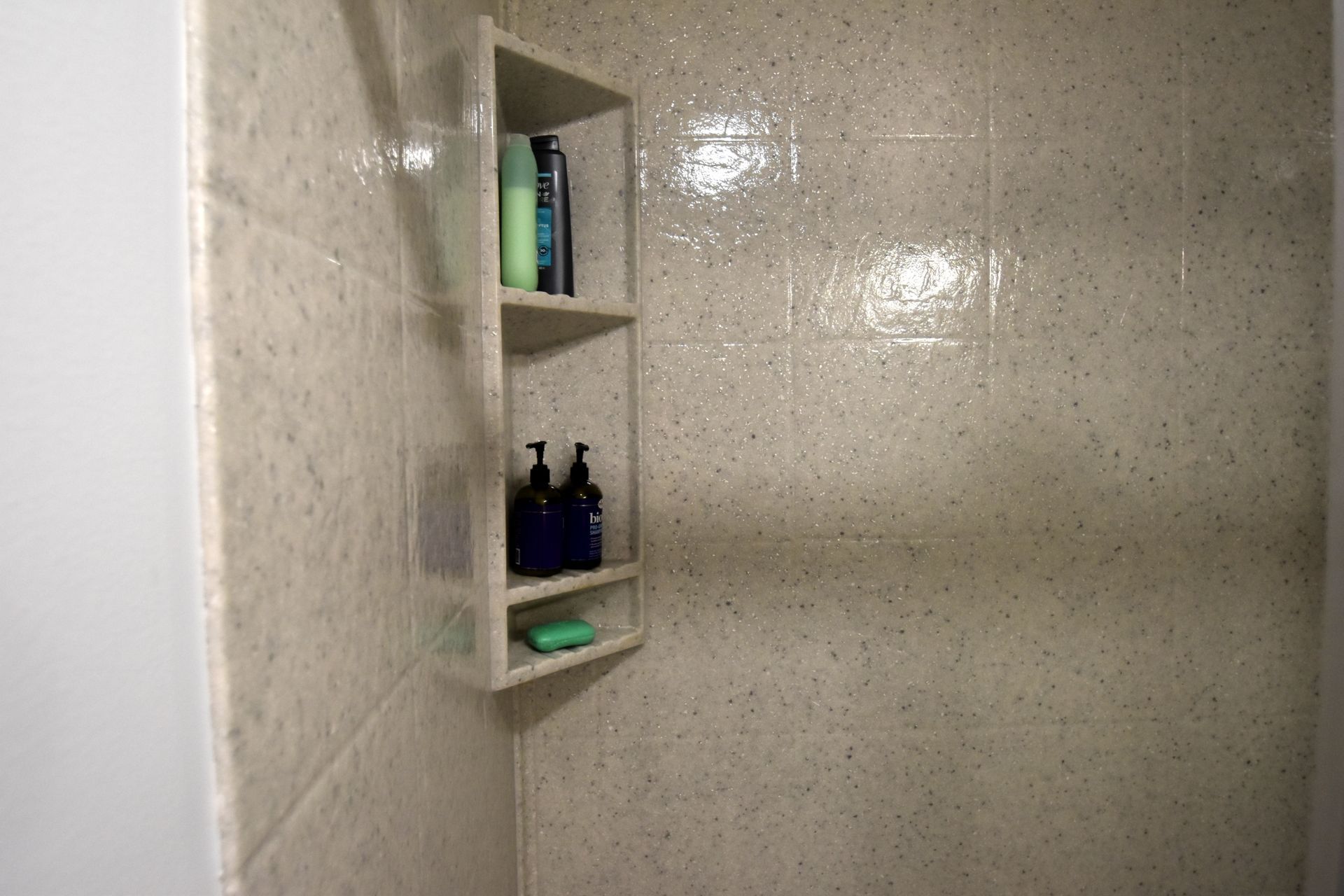 Corner shower shelf with toiletries, including bottles and a soap bar, against beige tile.