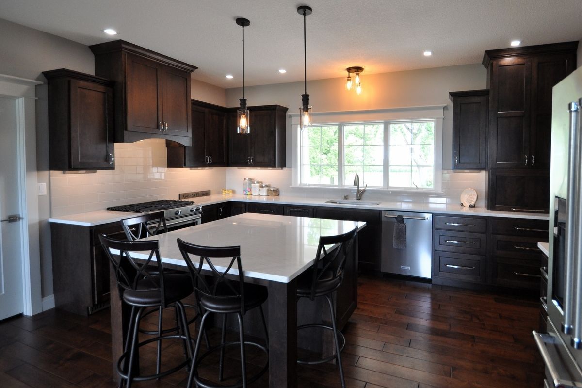 Modern kitchen with dark cabinets, white countertops, island stools, and hardwood floors, lit by pendant lights