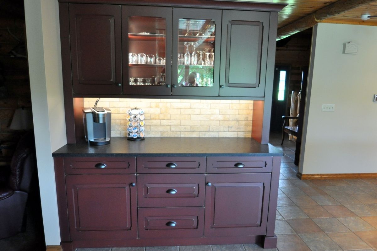Dark wood kitchen hutch with glass cabinets, countertop, and under-cabinet lighting against a tiled wall.