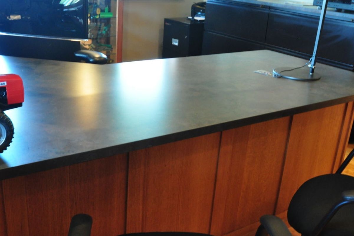 Wooden café counter with dark tabletop and metal railing, in a room with chairs and kitchen equipment.