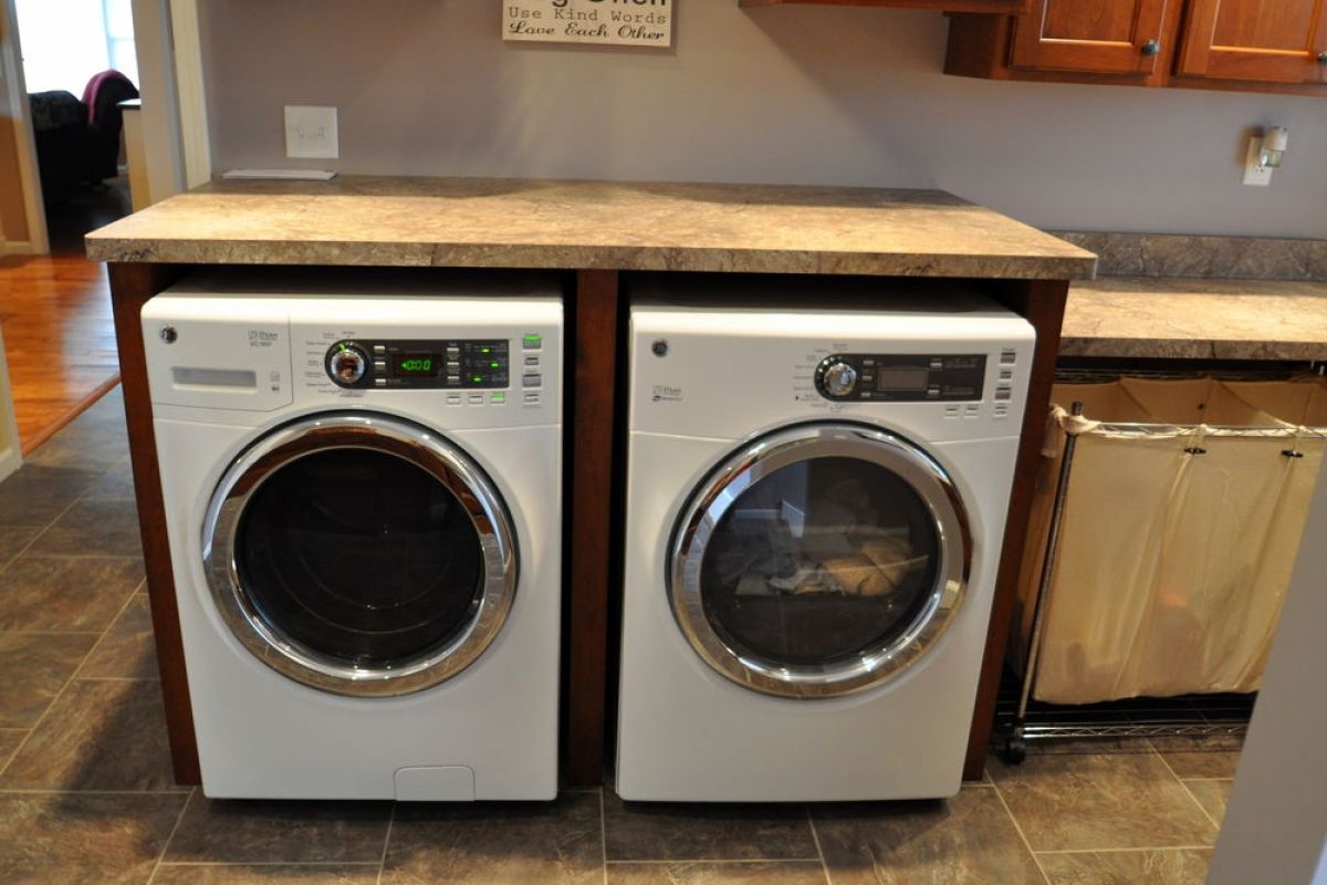 Two front-loading washers under a countertop in a kitchen with brown cabinets and tiled floor