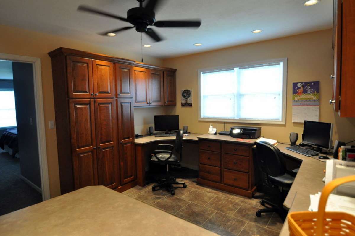 Home office with wooden cabinets, desks, computer monitors, and a ceiling fan by a window.