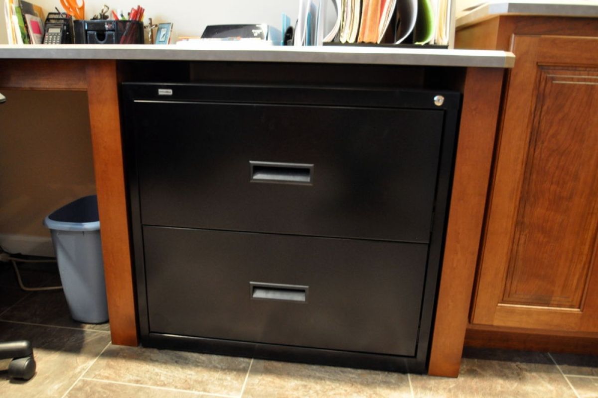 Black two-drawer cabinet beneath a kitchen counter, flanked by wood cabinets and a trash bin.