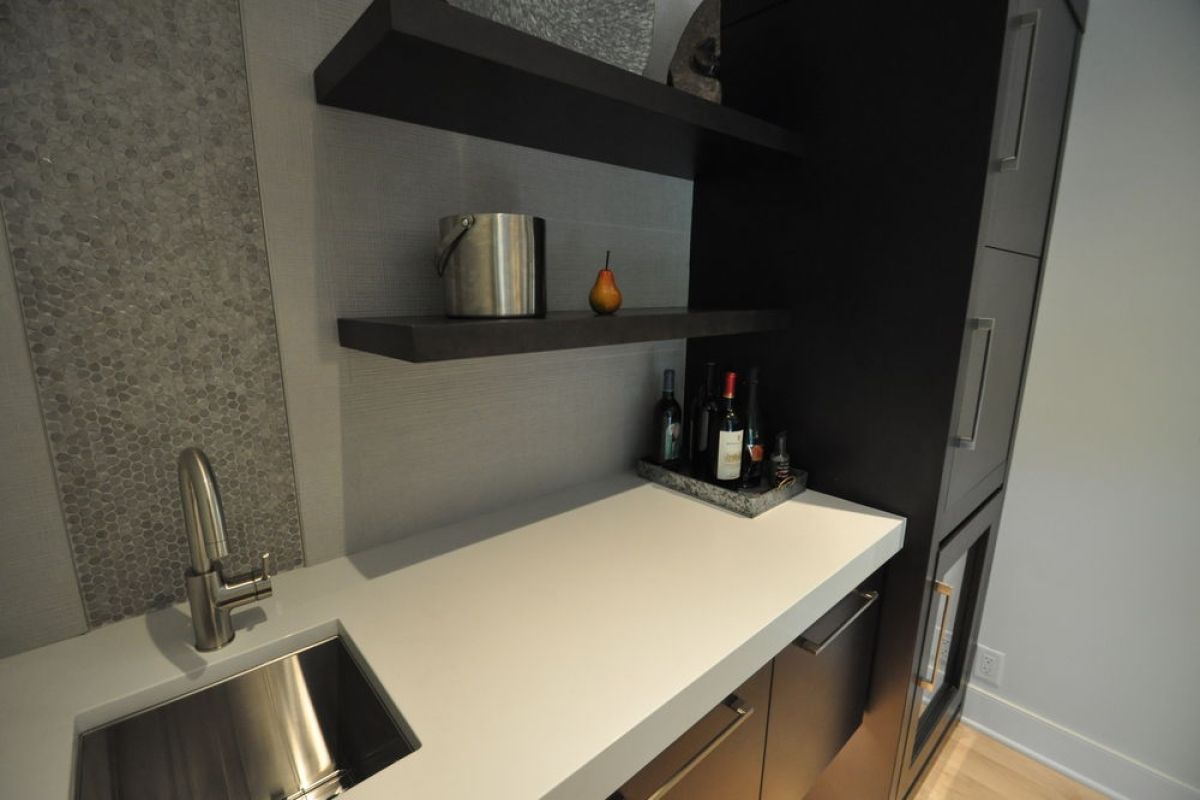 Modern kitchen counter with stainless sink, white countertop, dark shelves, and bottles by a window