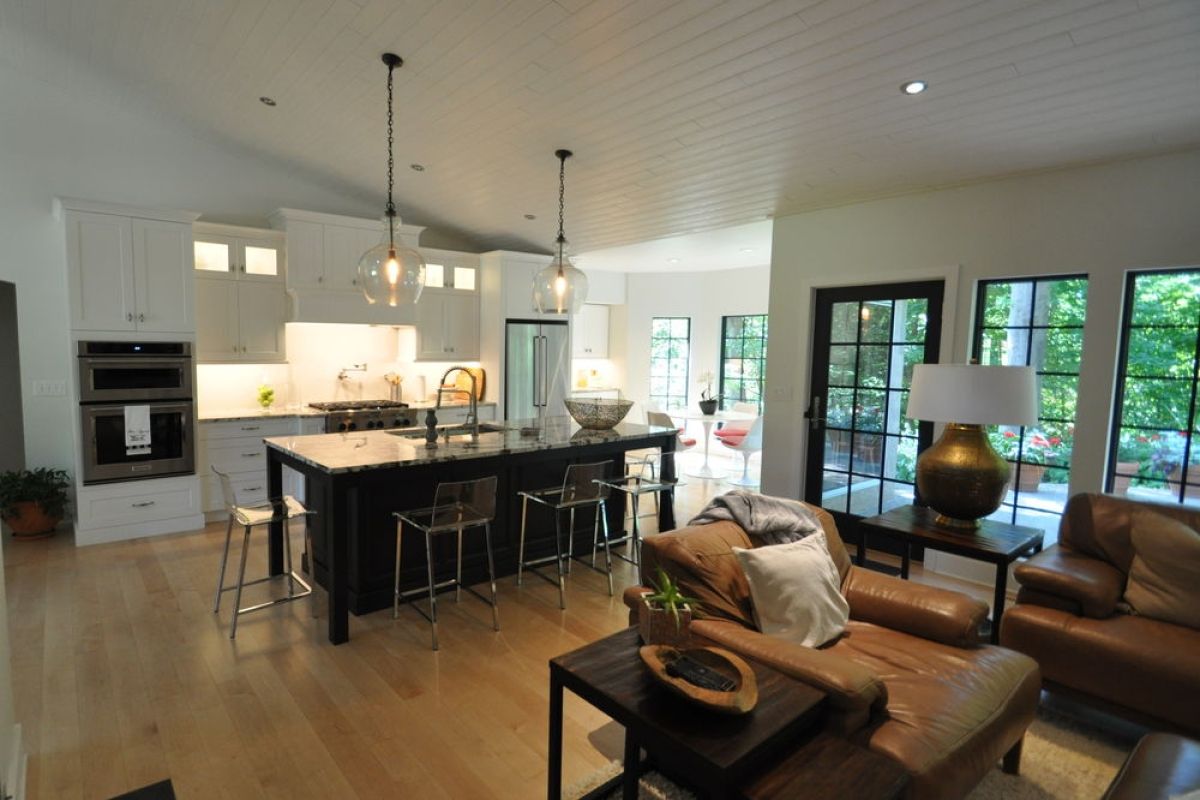 Open-concept kitchen and living room with dark island, leather chairs, and large windows overlooking greenery
