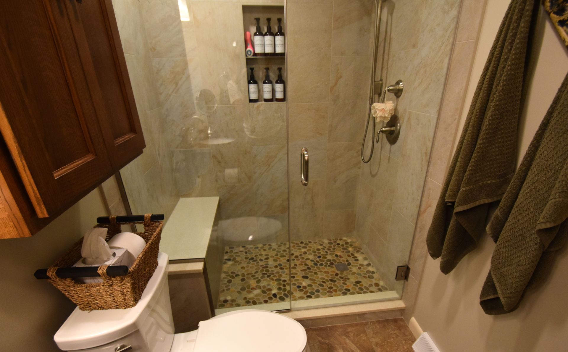 Small tiled bathroom with a glass shower, wall shelf, towel rack, and sink with basket of toiletries