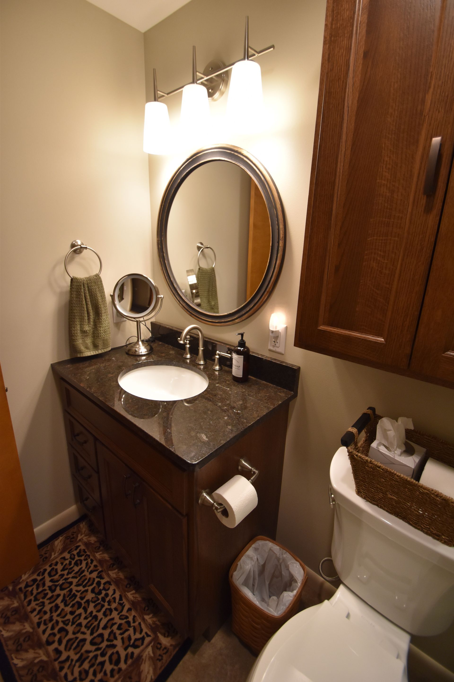 Small bathroom with dark wood vanity, oval mirror, bright light, toilet, and leopard-print rug.