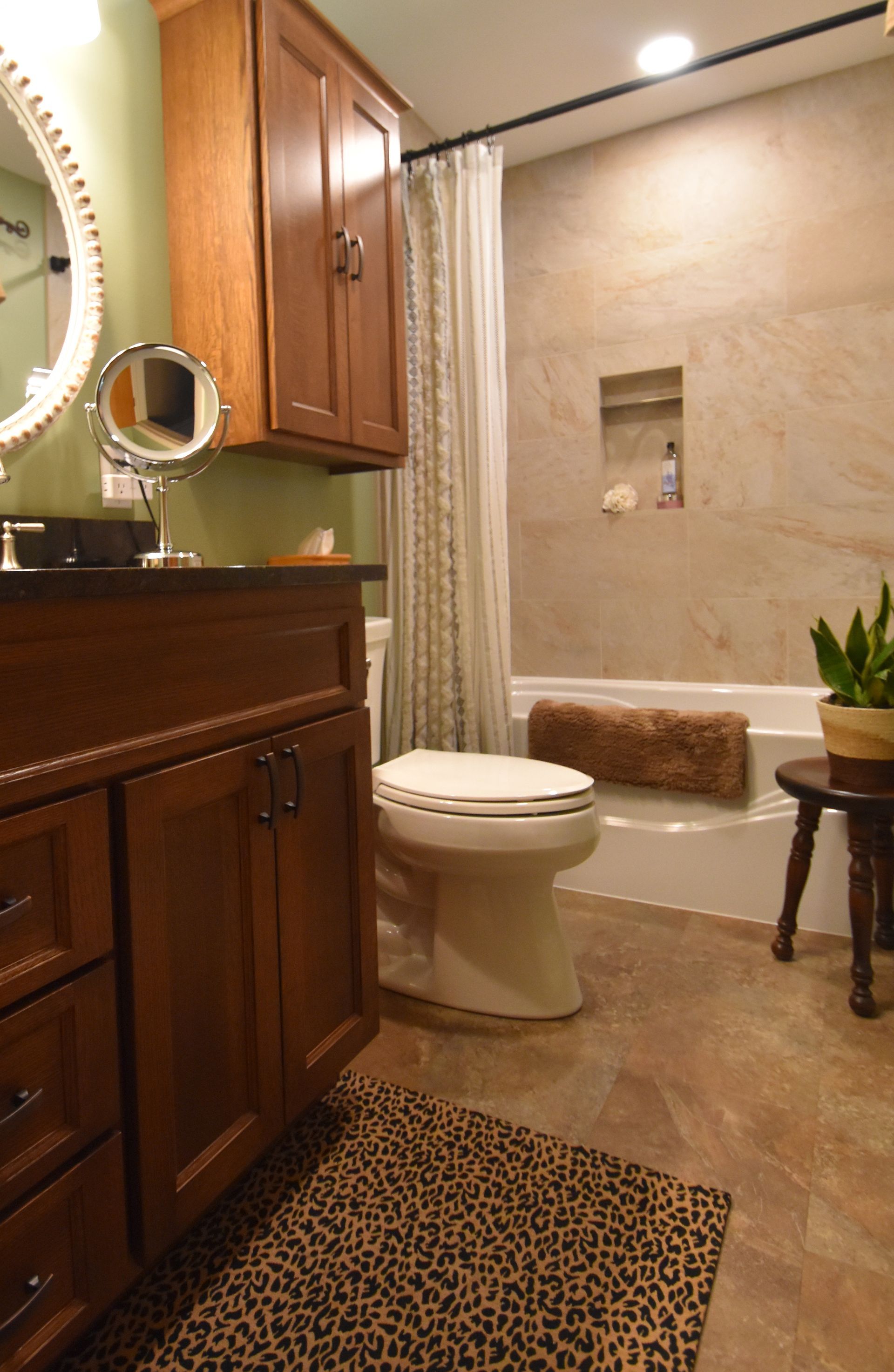 Small bathroom with wood vanity, toilet, tiled shower, and leopard-print rug.