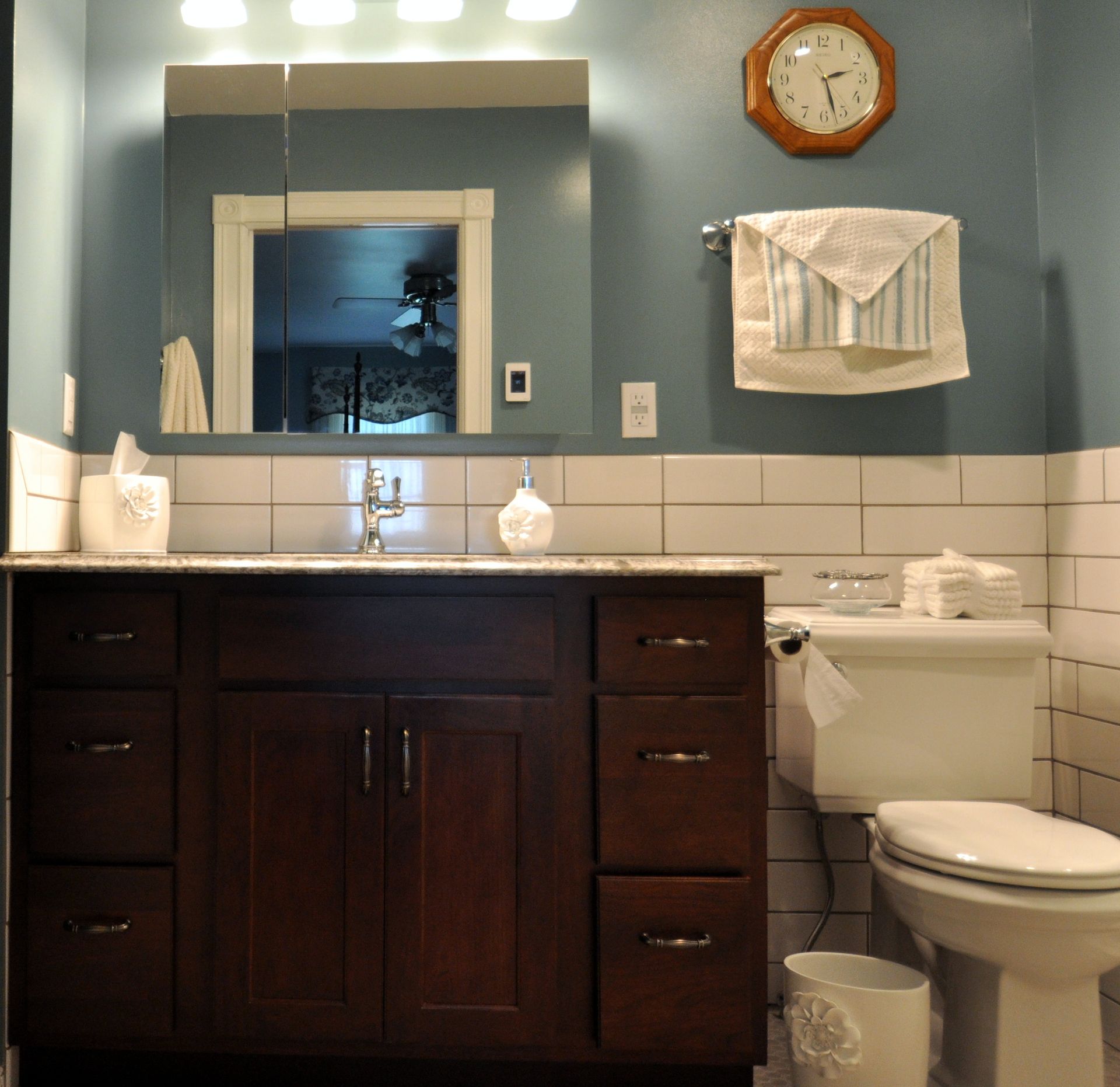 Bathroom vanity with dark wood cabinets, blue walls, mirror, sink, toilet, clock, and folded towels