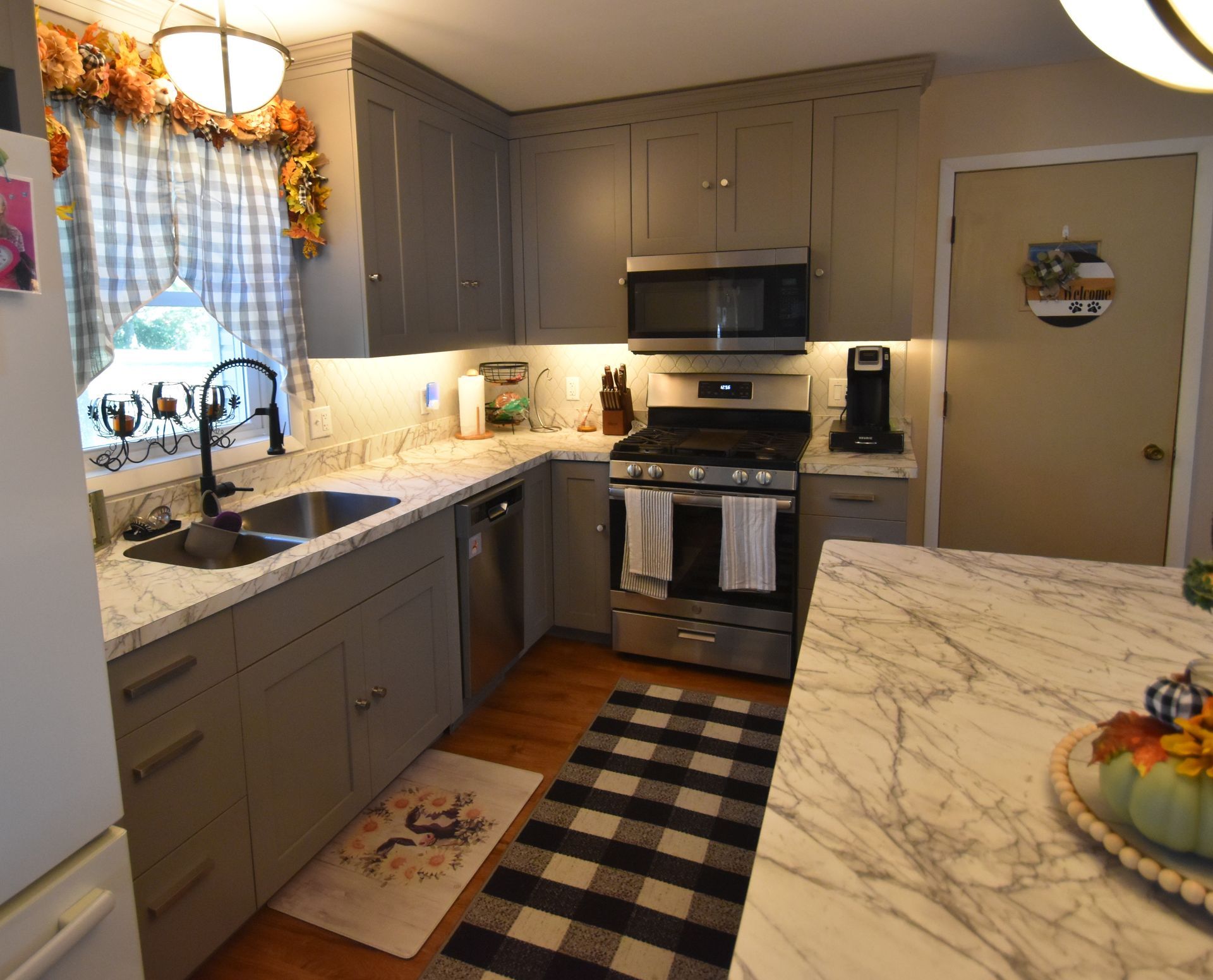 Small gray kitchen with marble counters, stainless appliances, and a black checkered rug near a beige door.
