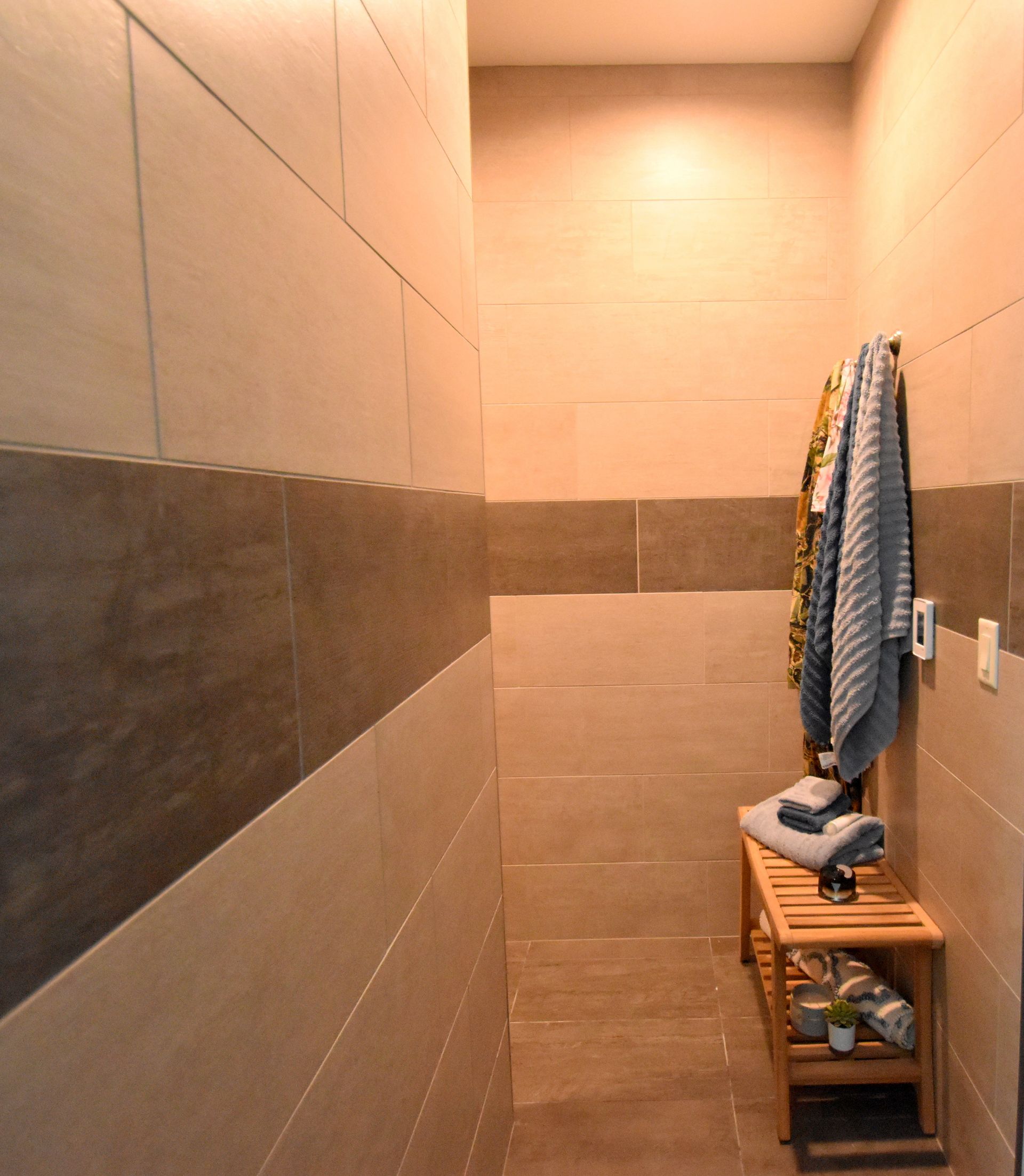 Narrow beige-tiled shower stall with a dark horizontal stripe and a small wooden shelf holding towels and toiletries