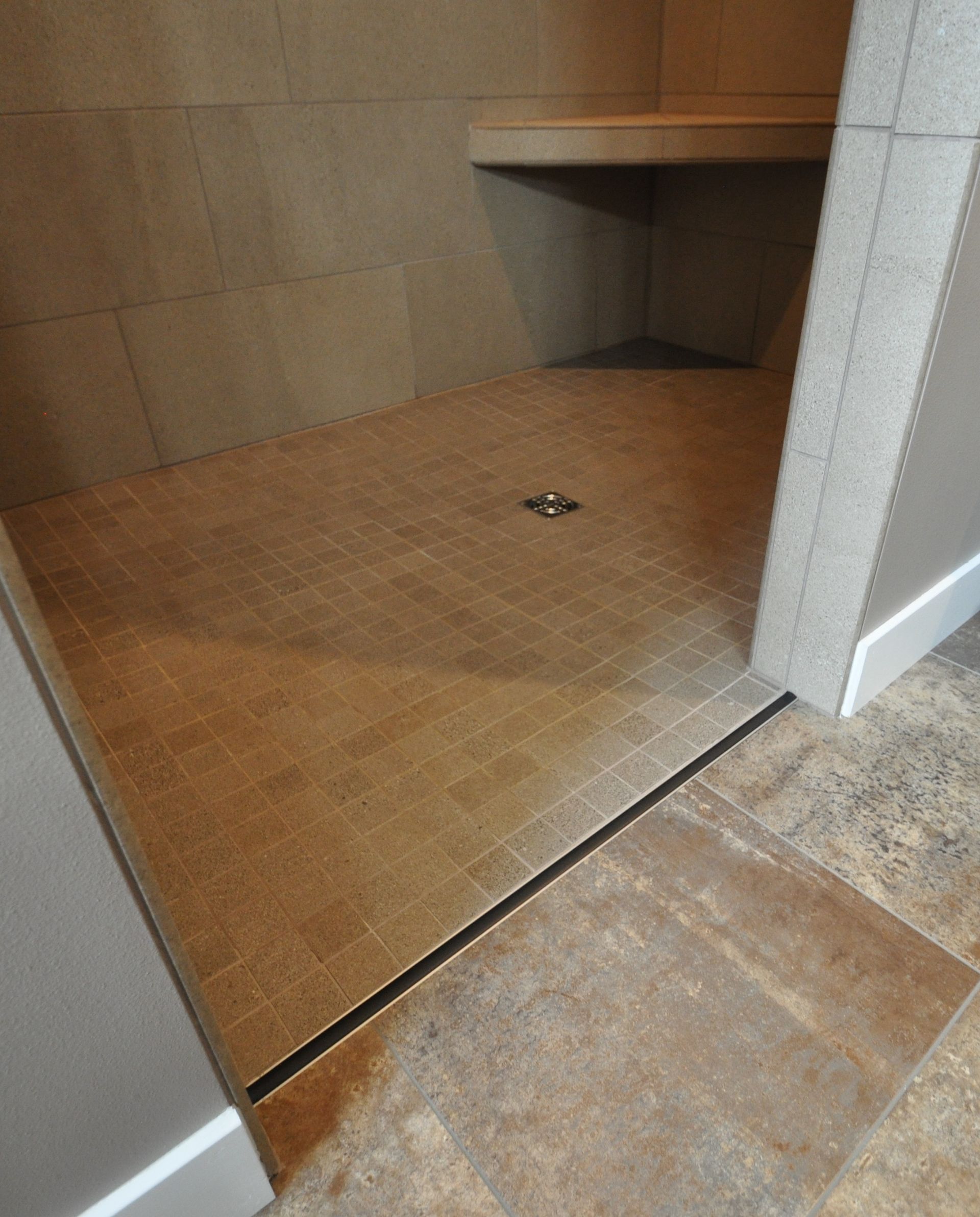 Small tiled shower stall with a wood-look floor and corner shelf, viewed through an open doorway