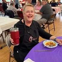 A woman is sitting at a table holding a red cup and smiling.