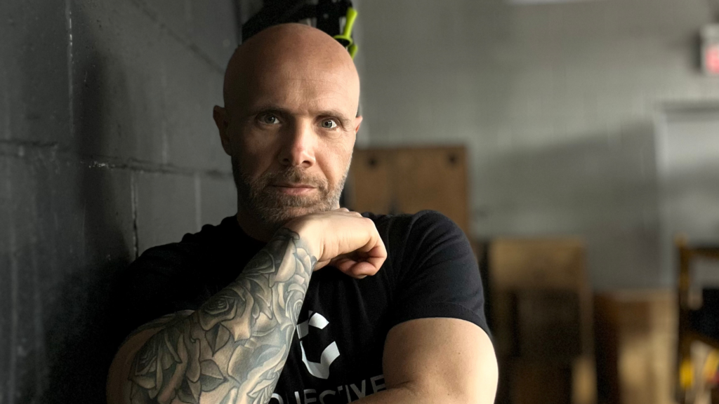 A bald man with a tattoo on his arm is leaning against a wall.