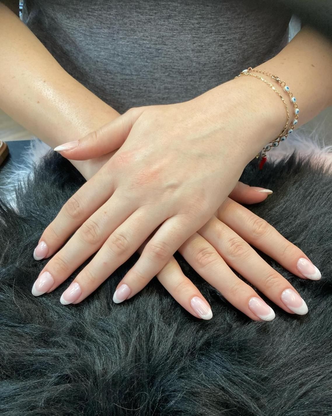 Hands with French tip nails, resting on a furry, gray surface. A gold bracelet adorns one wrist.